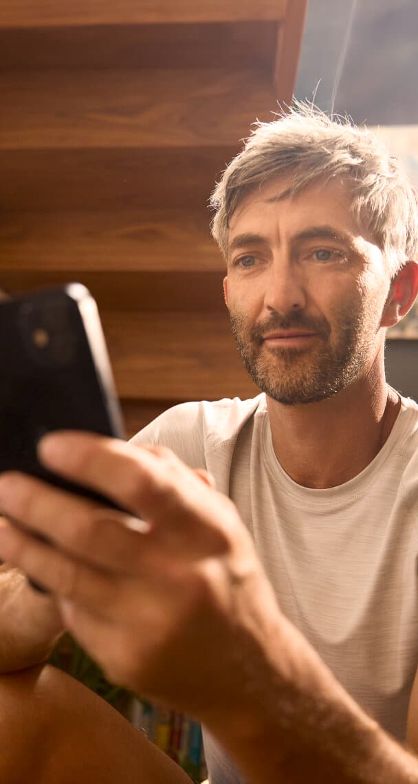 Ein Mann sitzt in einem Raum mit einer Treppe im Hintergrund. Er hält ein Smartphone in der linken Hand, auf das er schaut.