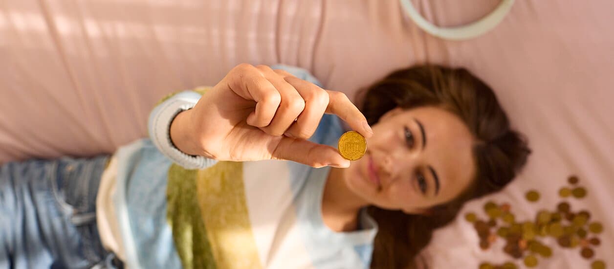 Eine Frau liegt auf einem weichen Untergrund, neben ihrem Kopf sind zahlreiche Münzen verstreut. Eine Münze hält sie nach oben, darauf liegt der Fokus.