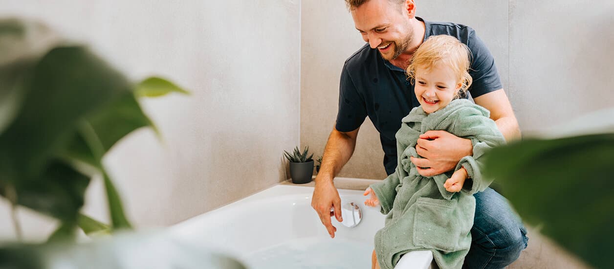 Ein Mann sitzt mit einem Kind im Arm auf einem Badewannenrand und wartet auf das einlaufende Wasser.