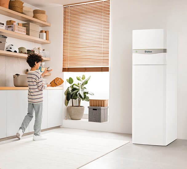 Ein Junge spielt in einem Raum mit Regalen links an der Wand und einer Wärmepumpe rechts im Bild mit einem Baseball und einem Baseballhandschuh.