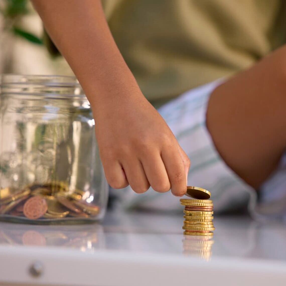 Eine Hand stapelt Geldmünzen aufeinander, im Hintergrund sieht man ein Glas mit Münzen und eine sitzende Person.
