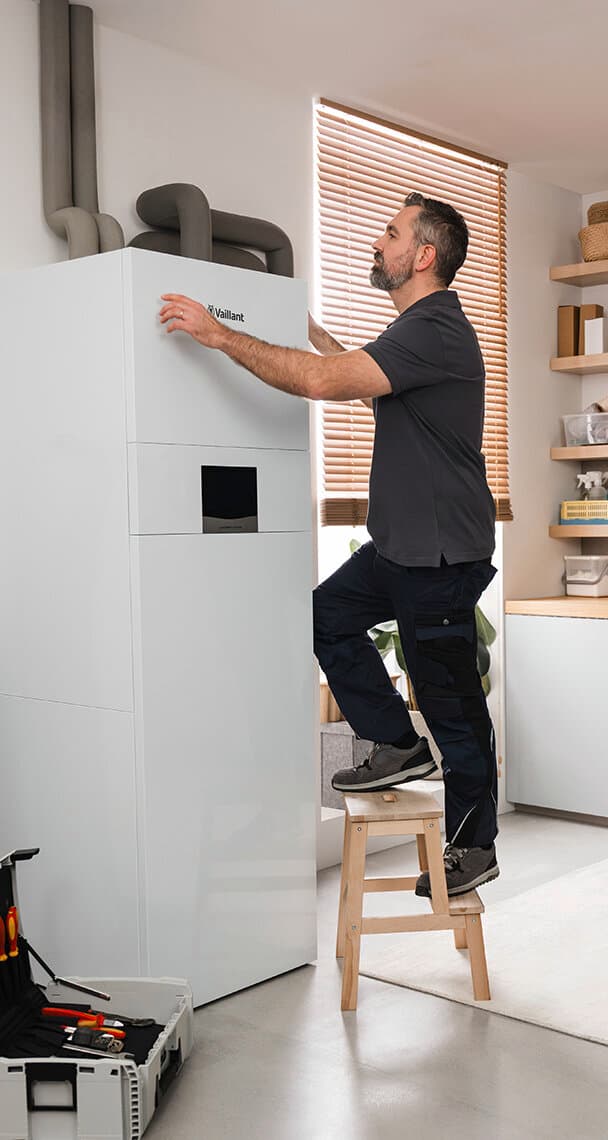 Ein Vaillant Fachhandwerker steht auf einem hölzernen zweistufigen Hocker und justiert mit den Händen eine Vaillant Erdwärmepumpe geoCOMPACT exclusive Inneneinheit, die in einem Raum mit Fenster und Regalen an der Wand steht.