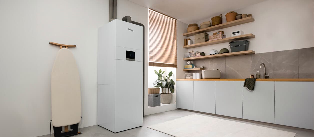 Eine Vaillant Erdwärmepumpe geoCOMPACT exclusive steht in einem Haushaltsraum zwischen einem an der Wand lehnenden Bügeleisen und einem bodenhohen Fenster. Rechts im Bild ist eine Art Küchenzeile mit Spülbecken zu sehen.