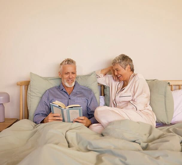 Ein Mann mit weißem Bart und weißen Haaren sitzt zugedeckt mit Kissen im Rücken in einem Bett, er hält ein aufgeschlagenes Buch in der Hand, das er anschaut. Neben ihm, ihm zugewandt, sitzt eine Frau im Pyjama mit grauen Haaren, die ihn anlächelt.