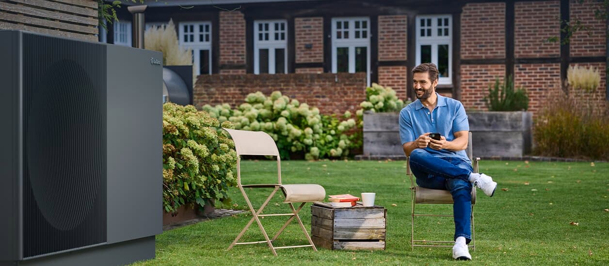 Eine Vaillant Wärmepumpe aroTHERM perform steht in einem gepflegten Garten neben einem Blumenbeet. Davor stehen auf dem Rasen zwei Klappstühle. Auf dem rechten Stuhl sitzt ein Mann mit einem Smartphone in der Hand.