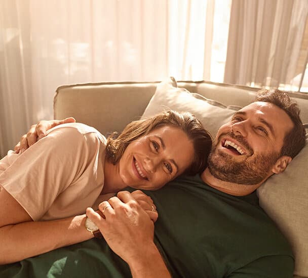 Ein Mann und eine Frau liegen Arm in Arm auf einer Couch. Im Hintergrund sieht man Gardinen-Vorhänge vor einem Fenster. Die Frau liegt auf der Brust des Mannes und die beiden halten sich an der Hand.