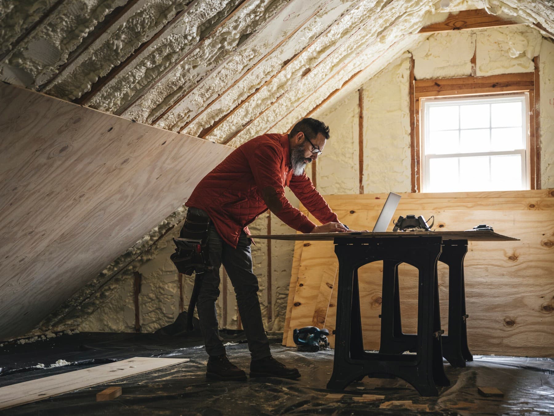 Handwerker, der während der Isolierung eines lichtdurchfluteten Dachgeschosses auf einen Laptop schaut