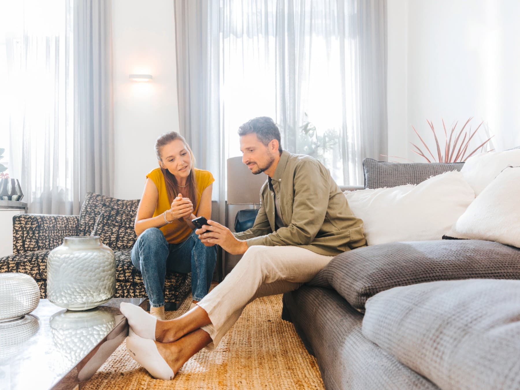 Ein Mann und eine Frau sitzen auf einer großen Couch in einem Wohnzimmer mit Couch, Beistelltisch und Regalen. Sie schauen gemeinsam auf ein Smartphone.