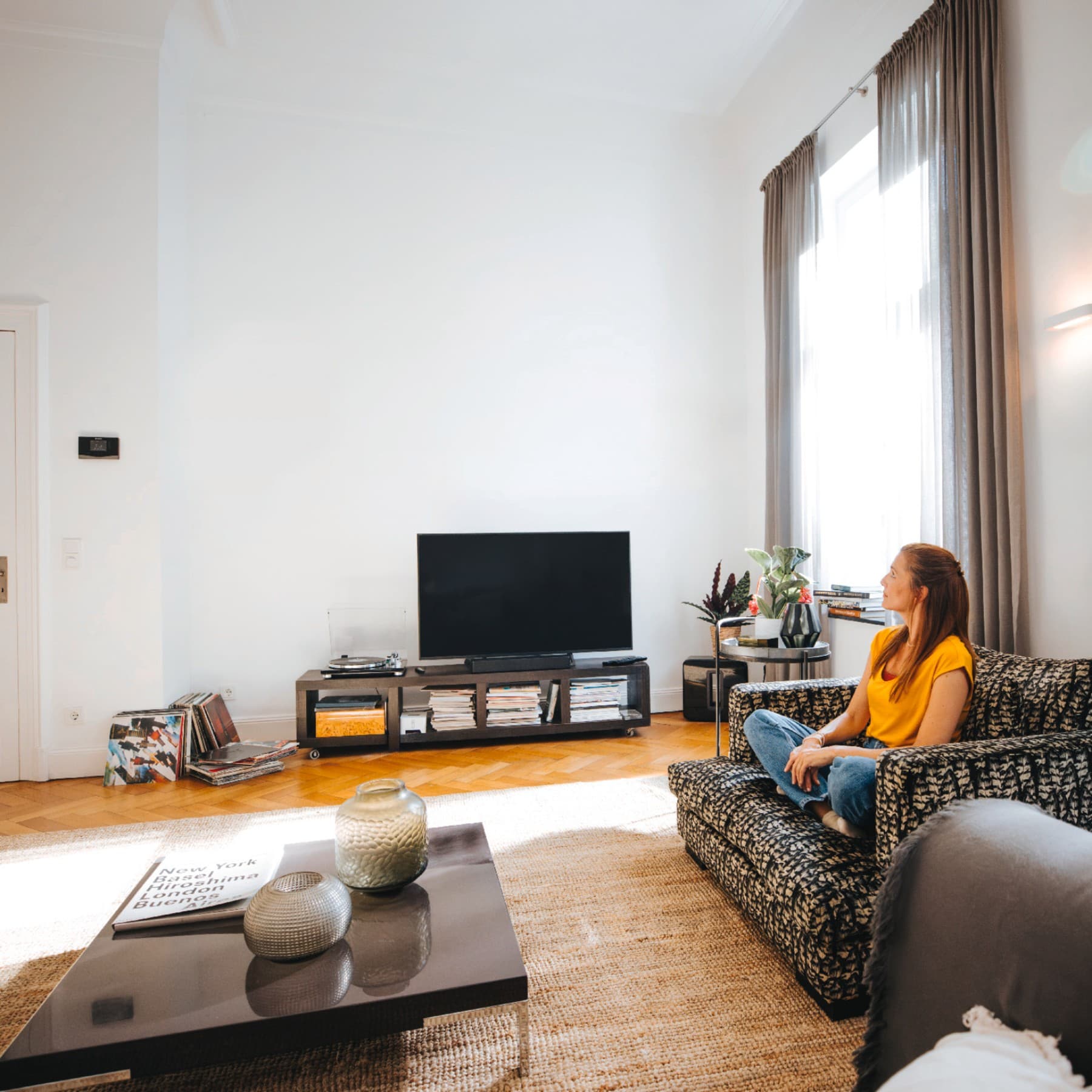 Eine Frau sitzt auf einem Wohnzimmer in einem hellen, offenen Wohnzimmer, das mit Couch, Tisch, Fernsehtisch und Fernseher eingerichtet ist. Sie schaut zu einer Tür, neben dem ein Vaillant Regler hängt.