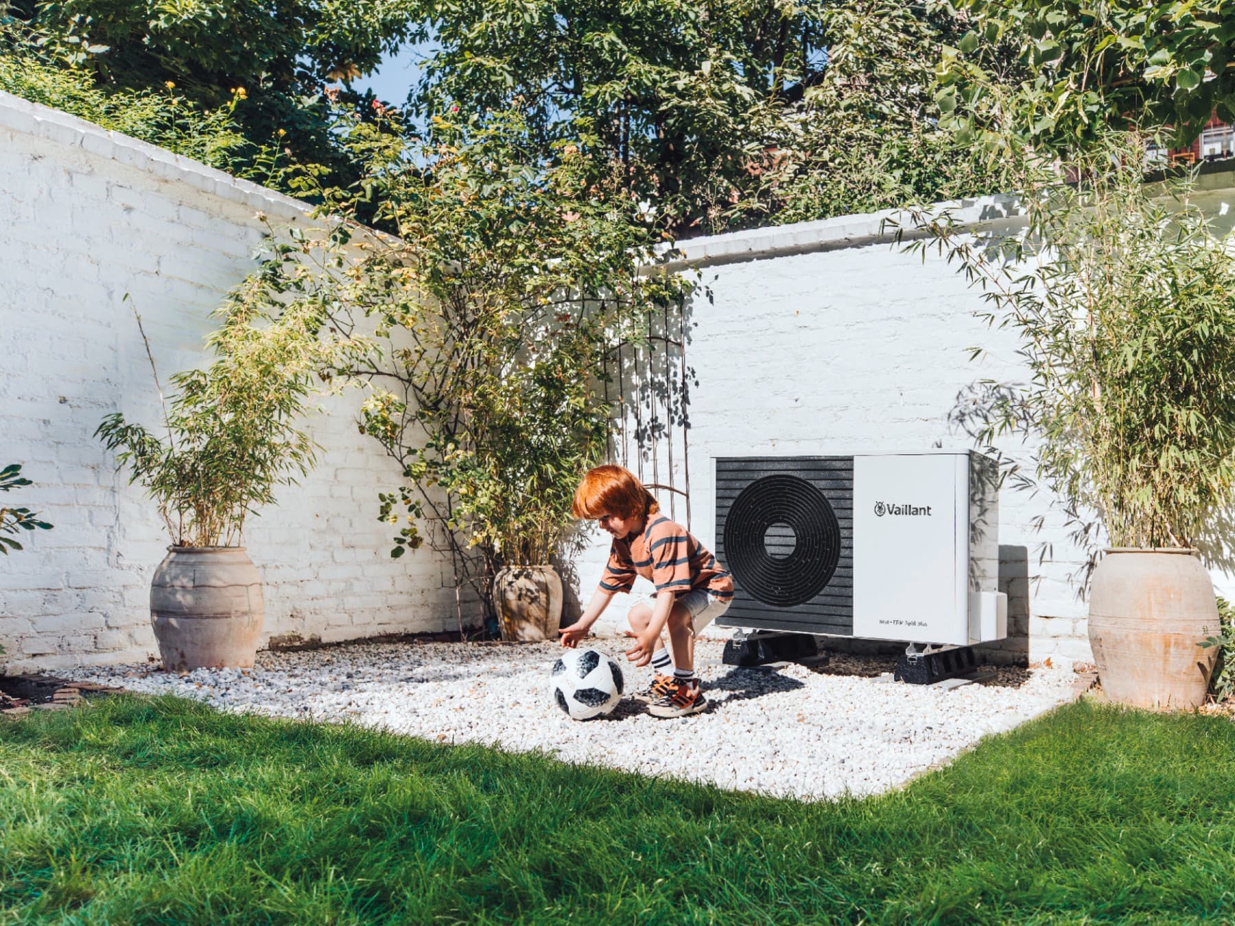 Kind spielt im Garten mit einem Ball vor einer Split-Wärmepumpe aroTHERM Split plus von Vaillant