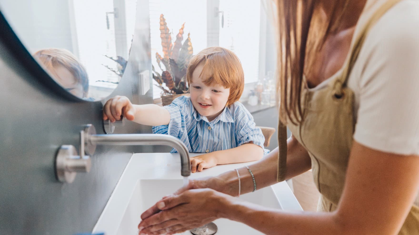 Eine Frau wäscht sich am Waschbecken die Hände. Der Wasserhahn wird von einem Kind im Hintergrund bedient.