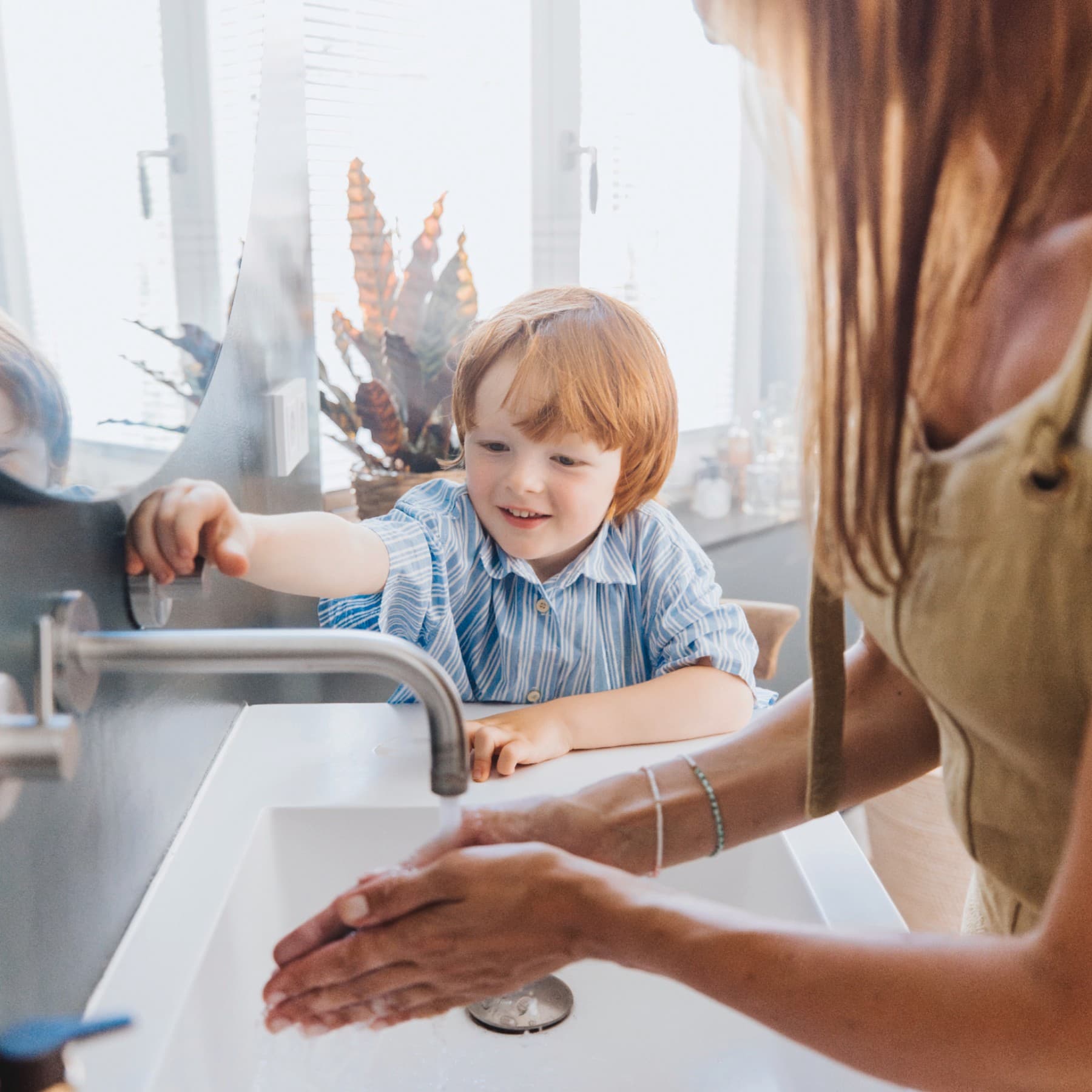 Eine Frau wäscht sich an einem Waschbecken die Hände, während ein Kind an der Seite steht und das Wasser anmacht.