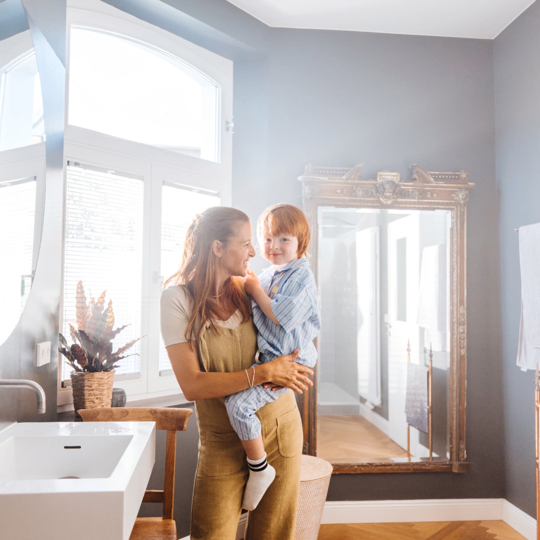 Eine Frau steht mit einem Kind auf dem Arm in einem hellen Badezimmer am Waschbecken und schaut das Kind an.