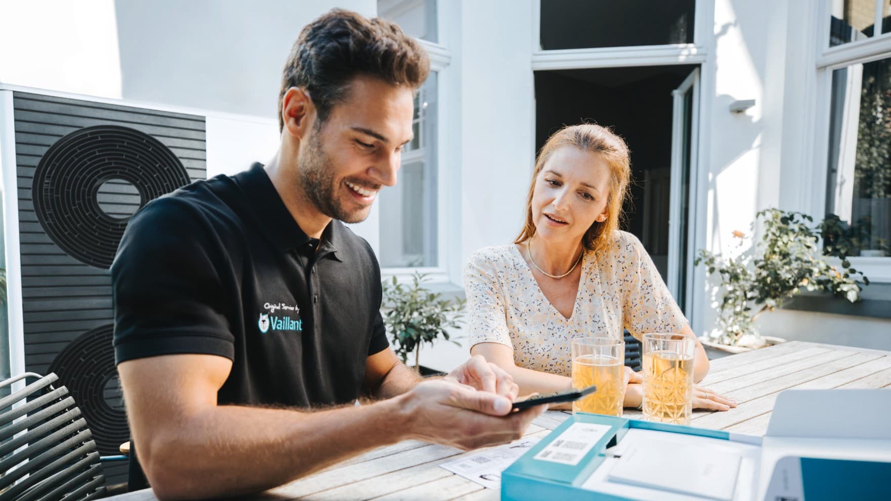 Ein Vaillant Fachhandwerker sitzt mit einer Kundin an einem Gartentisch, vor ihnen stehen zwei Gläser mit Getränken. Der Fachhandwerker zeigt der Kundin etwas auf einem Smartphone. Im Hintergrund ist eine Hausfassade mit Terrassentür sowie eine Vaillant Wärmepumpe zu sehen.
