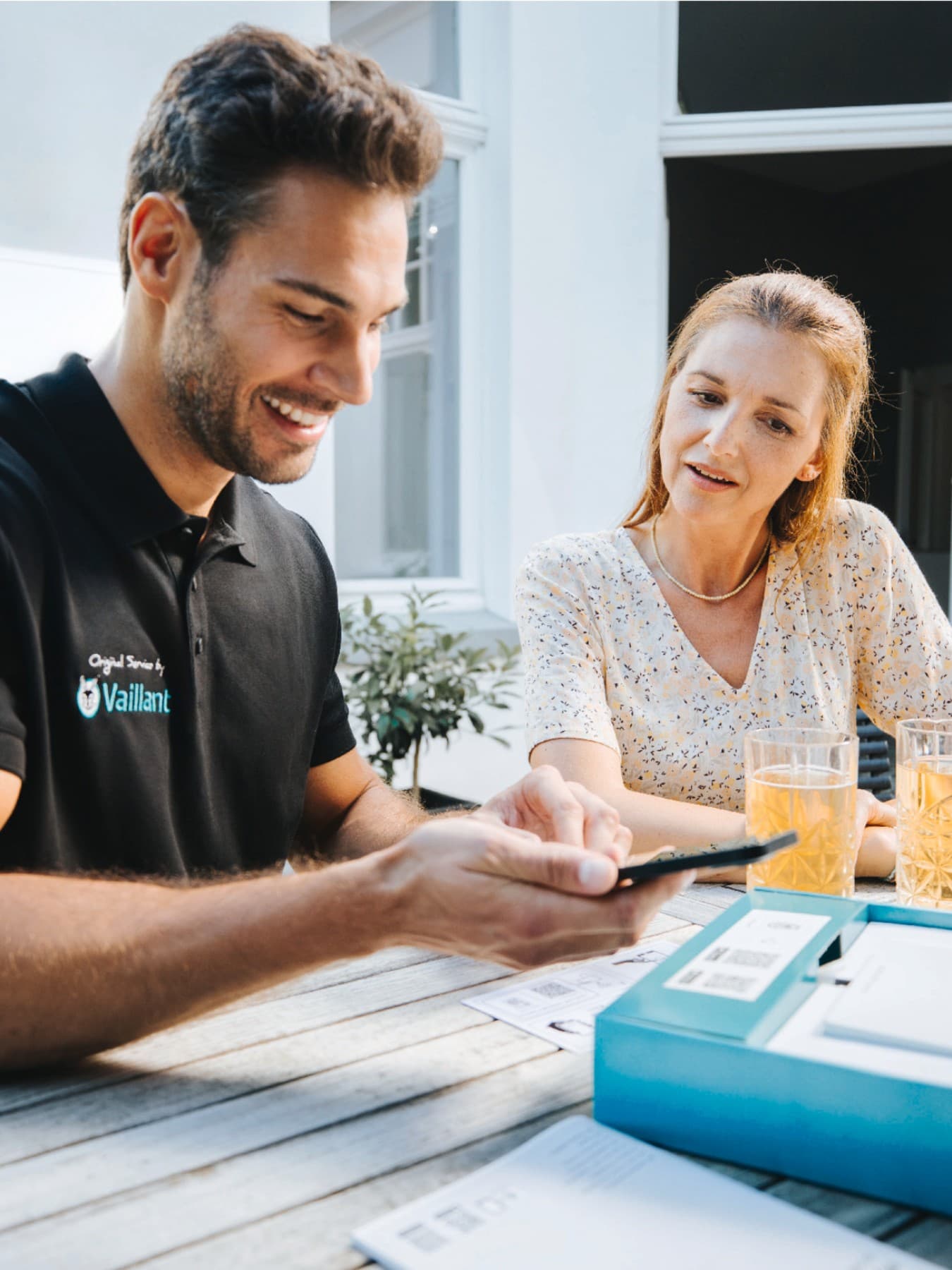 Ein Vaillant Fachhandwerker sitzt mit einer Kundin an einem Gartentisch, vor ihnen stehen zwei Gläser mit Getränken. Der Fachhandwerker zeigt der Kundin etwas auf einem Smartphone. Im Hintergrund ist eine Hausfassade mit Terrassentür sowie eine Vaillant Wärmepumpe zu sehen.