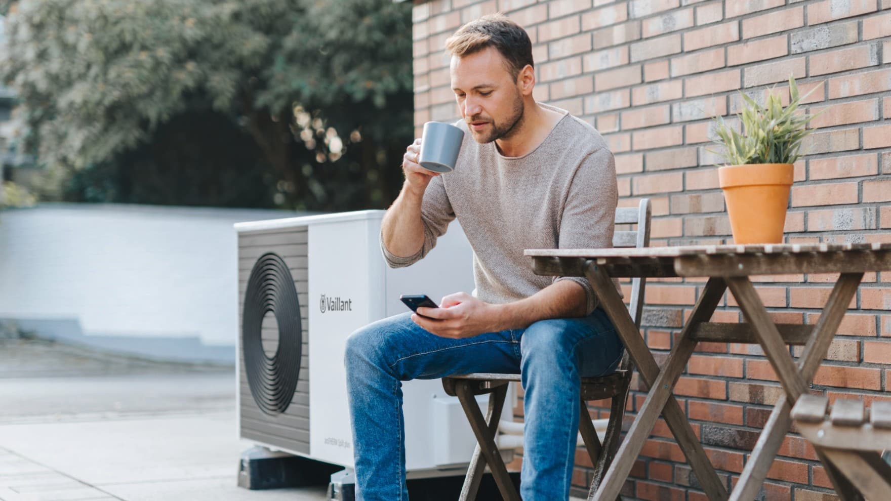Mann sitzt mit einer Tasse Kaffee und Handy vor dem Haus nehmen einer Split-Wärmepumpe aroTHERM Split plus von Vaillant