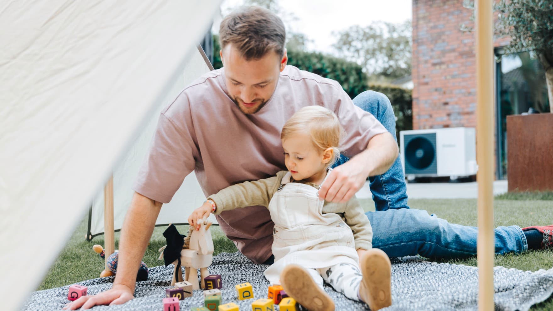 Ein Mann spielt mit einem Kind auf einer Decke im Garten mit Bauklötzen. Im Hintergrund ist eine Vaillant Wärmepumpe zu sehen.