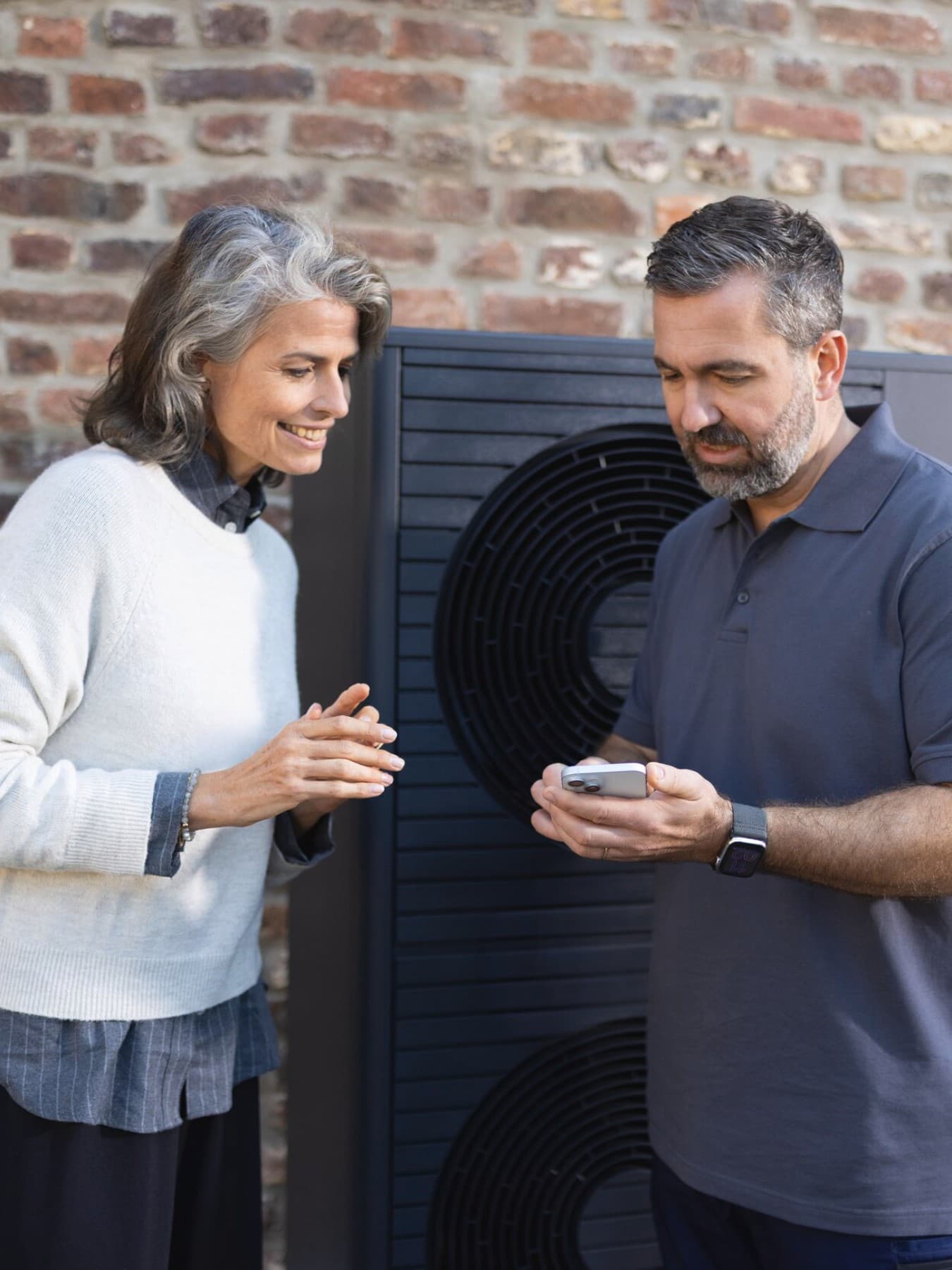 Ein Vaillant Fachhandwerker steht mit einer Kundin vor einer Vaillant Wärmepumpe, die vor einem Backsteingebäude steht. Der Fachhandwerker zeigt der Kundin etwas auf seinem Smartphone.