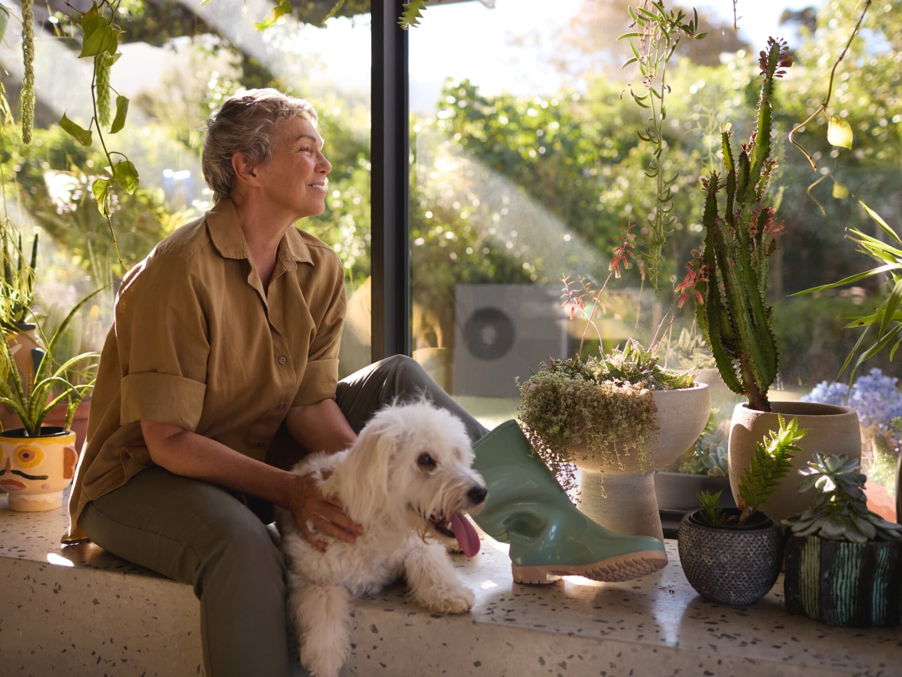 Eine Frau sitzt mit einem weißen Hund zwischen Topfpflanzen. Im Hintergrund sieht man eine schwarzgraue Vaillant Wärmepumpe.