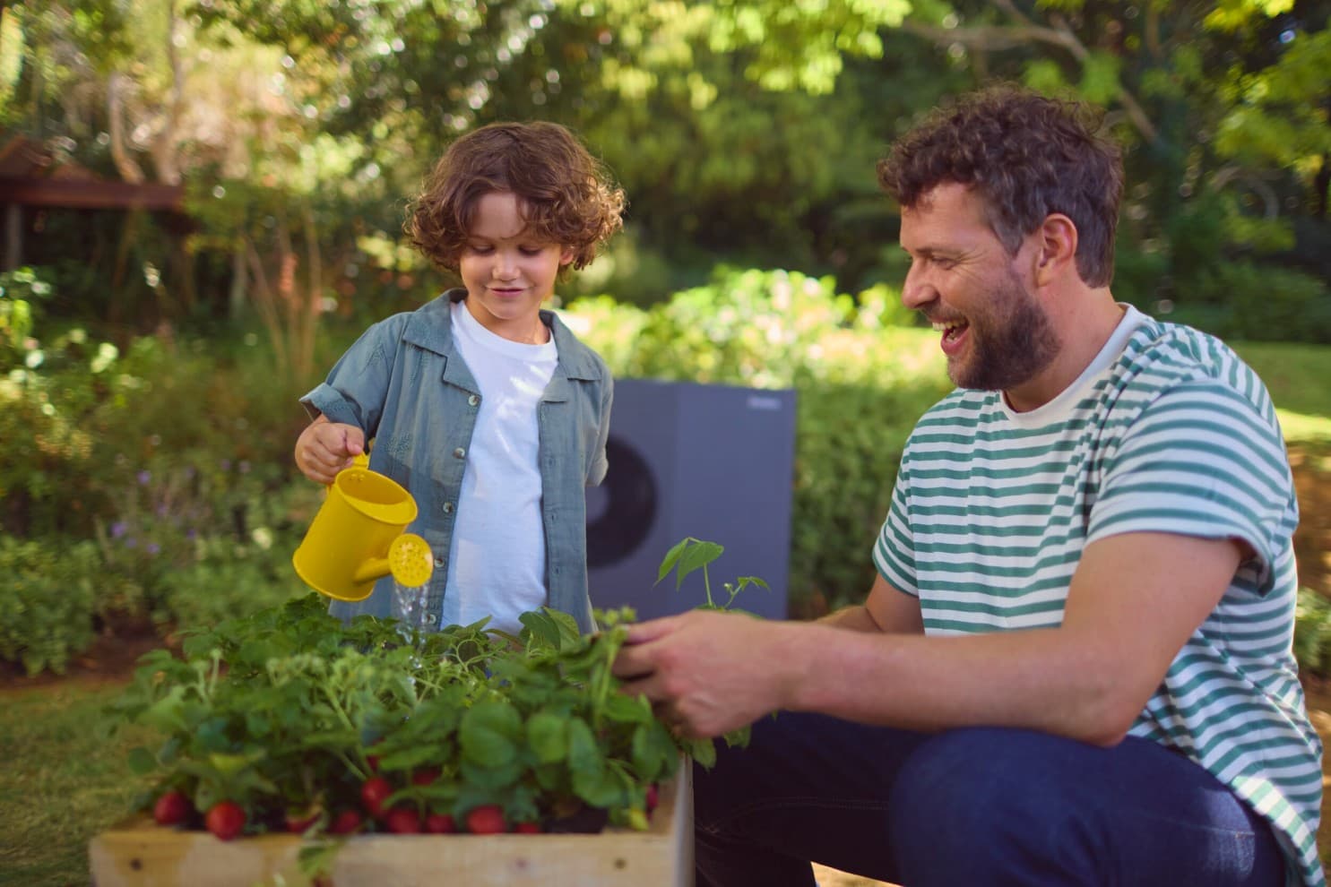 Vater und Sohn im Garten. Sohn gießt mit einer gelben Gießkanne die Pflanzen. Im Hintergrund sieht man eine aroTHEM plus Wärmepumpe