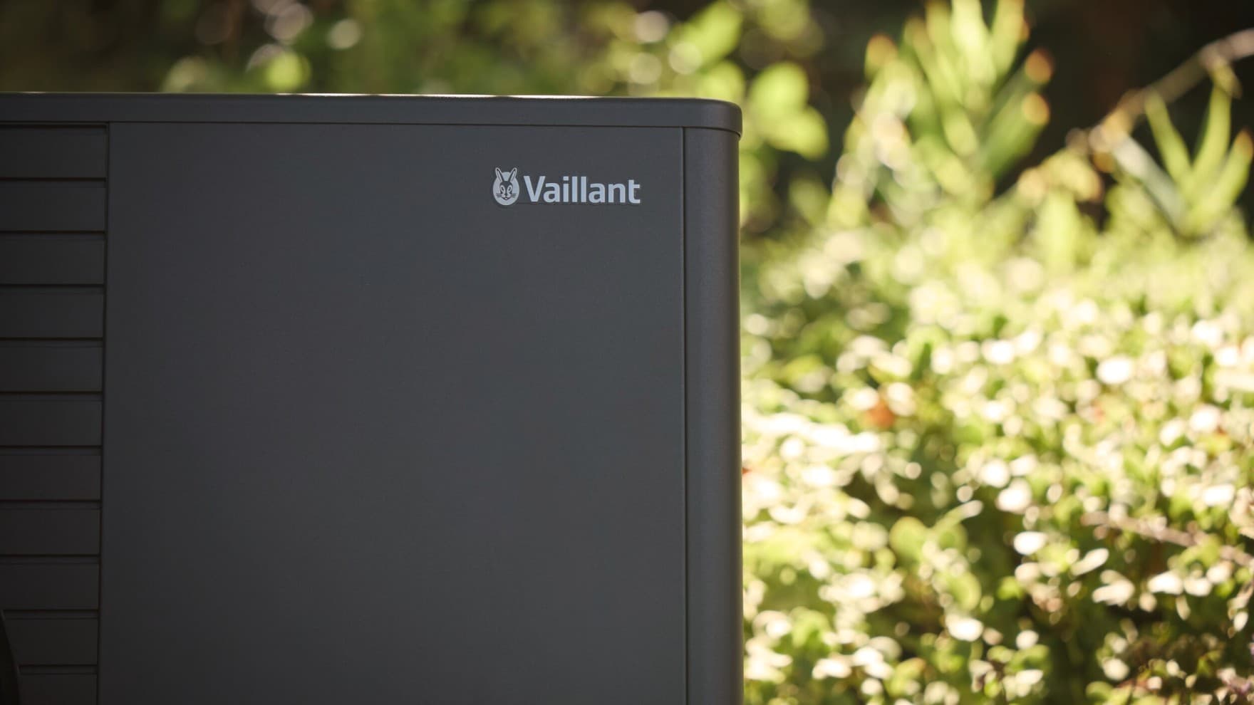 Eine Nahaufnahme einer Vaillant Wärmepumpe mit dem Fokus auf dem Vaillant Logo in der oberen rechten Ecke, im Hintergrund sieht man eine von Sonne beschienene Hecke.