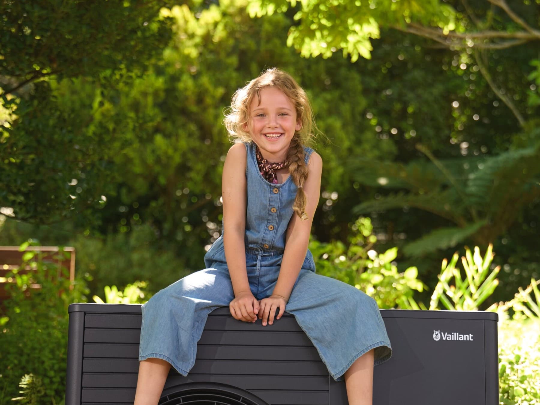 Lachendes Mädchen sitzt auf der dunklen Vaillant Wärmepumpe aroTHERM plus im sommerlchen Garten.