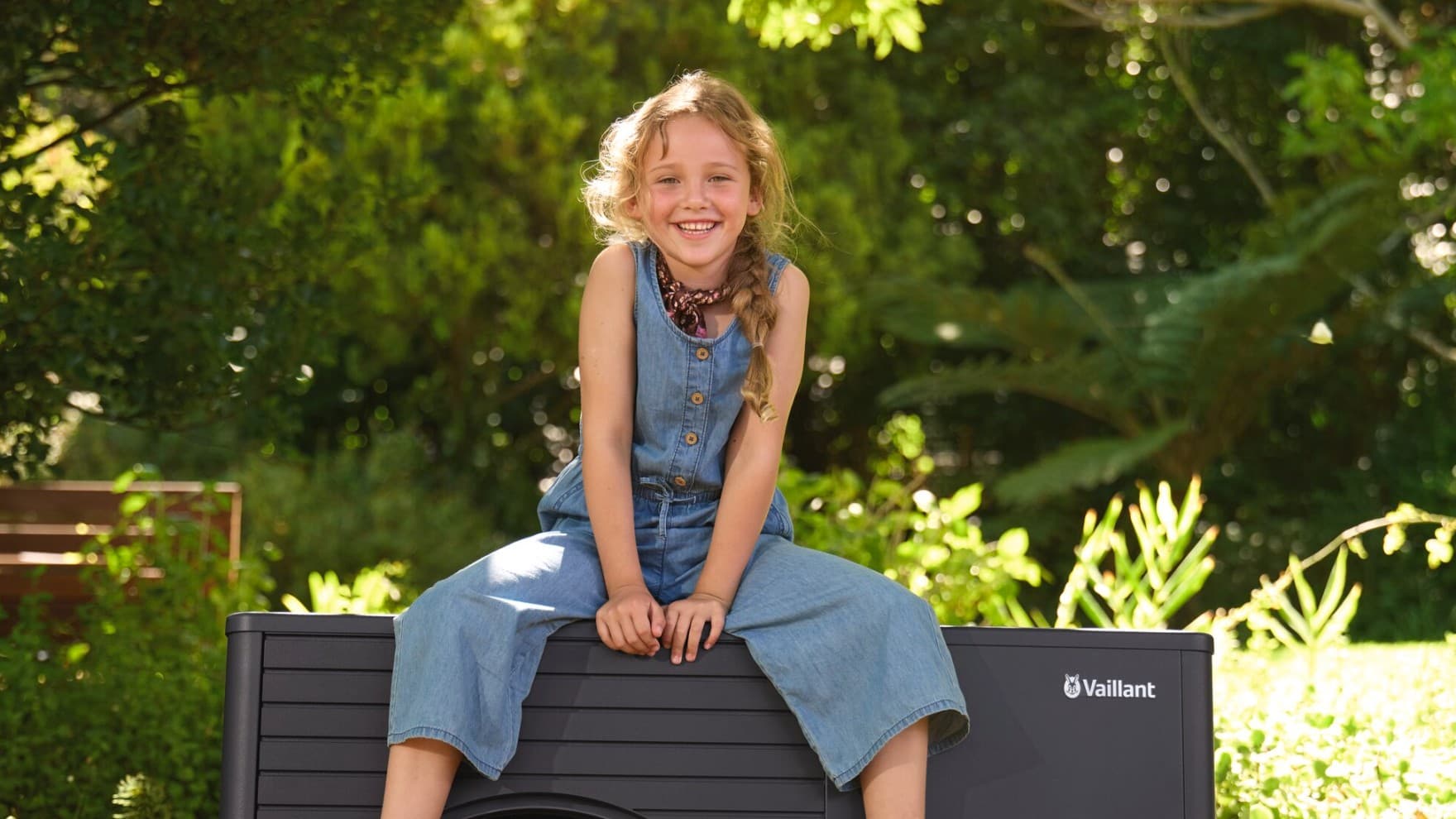 Kleines, lächelndes Mädchen sitzt auf einer aroTHERM plus Wärmepumpe im Garten mit Bäumen im Hintergrund