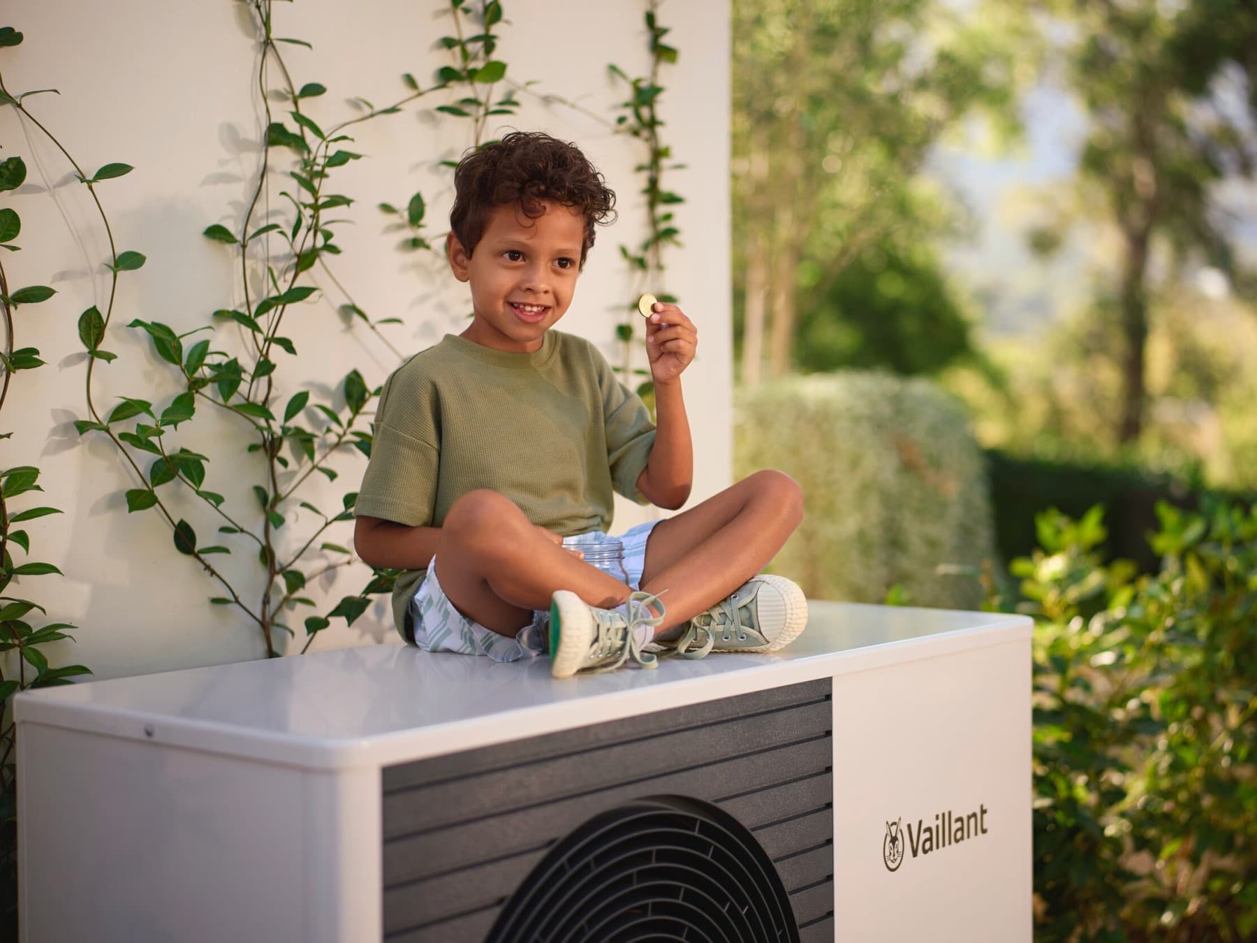 Junge, der eine Münze in der Hand hält und auf einer Außeneinheit einer Vaillant Wärmepumpe sitzt