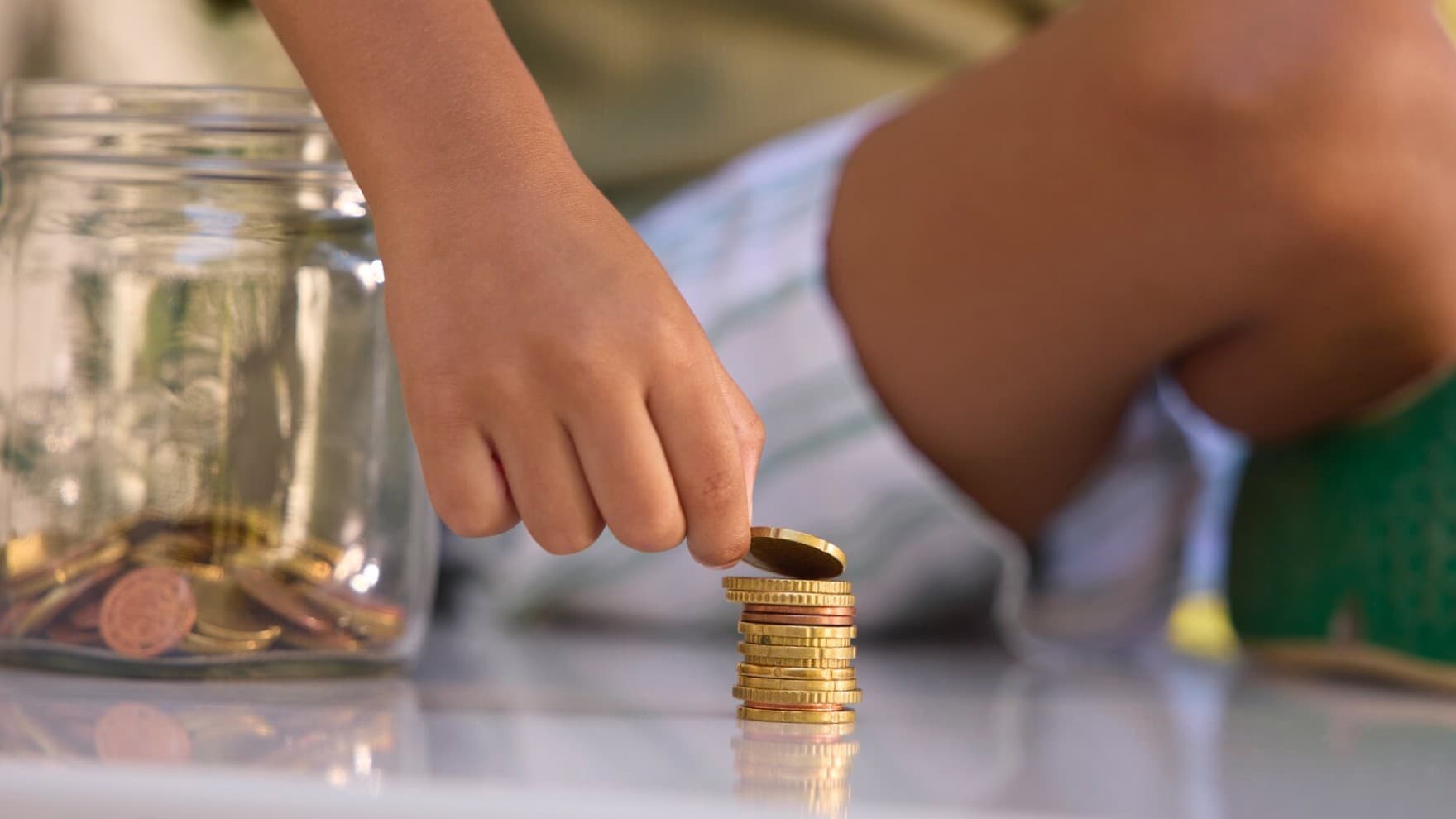 Eine Kinderhand stapelt Münzen neben einem Glas voller Münzen.