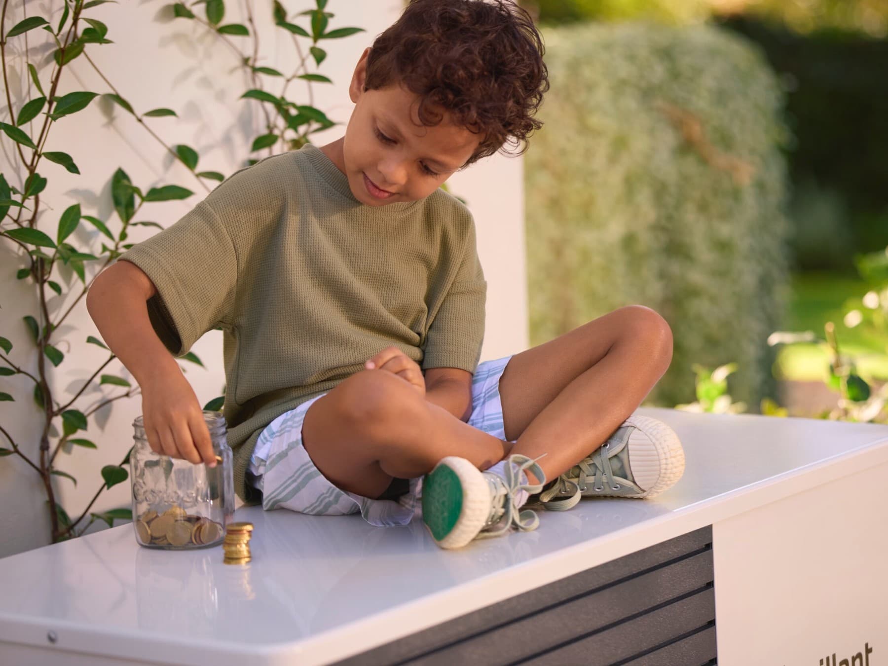 Ein Junge sitzt im Schneidersitz auf einer Vaillant Wärmepumpe und stapelt Geldmünzen neben einem Einmachglas.