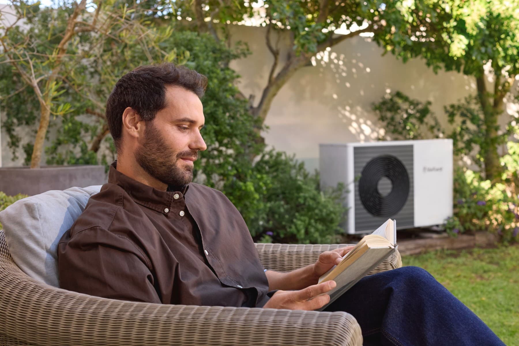 Mann sitzt im Garten und ließt ein Buch. Im Hintergrund steht eine Wärmepumpe von Vaillant.