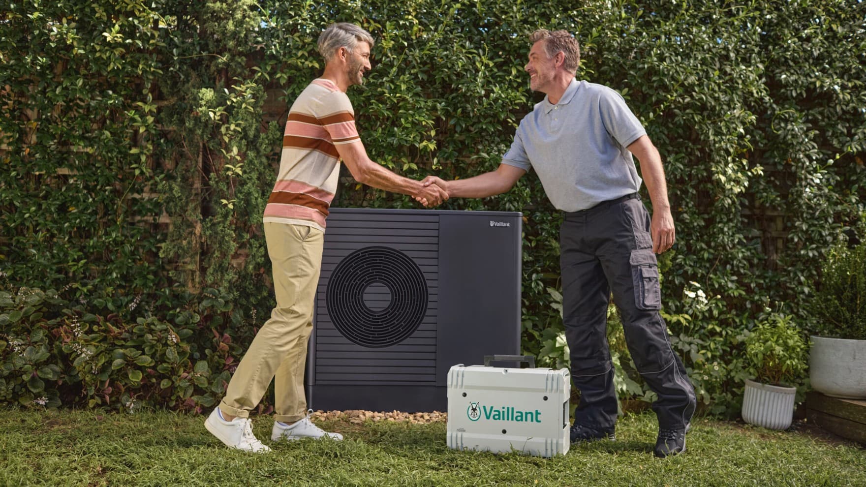 Ein Vaillant Fachpartner steht rechts im Bild und schüttelt einem Kunden die Hand. Sie stehen dabei im Garten vor einer Vaillant Wärmepumpe, zwischen ihnen steht ein Vaillant Werkzeugkoffer.
