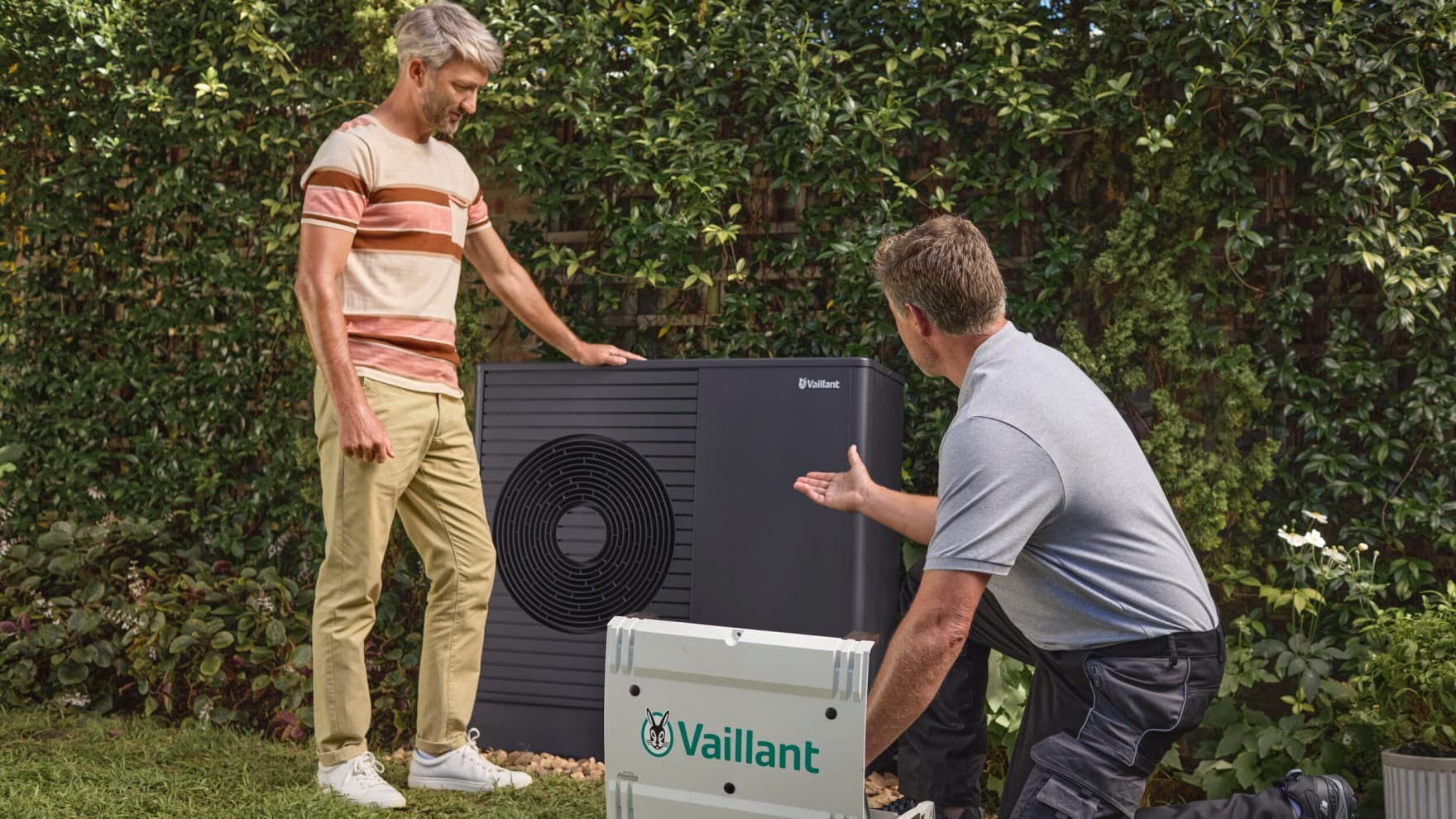Ein Vaillant Techniker kniet rechts im Bild und erklärt einem Kunden links im Bild die Wartung einer Wärmepumpe, die hinter ihnen steht.