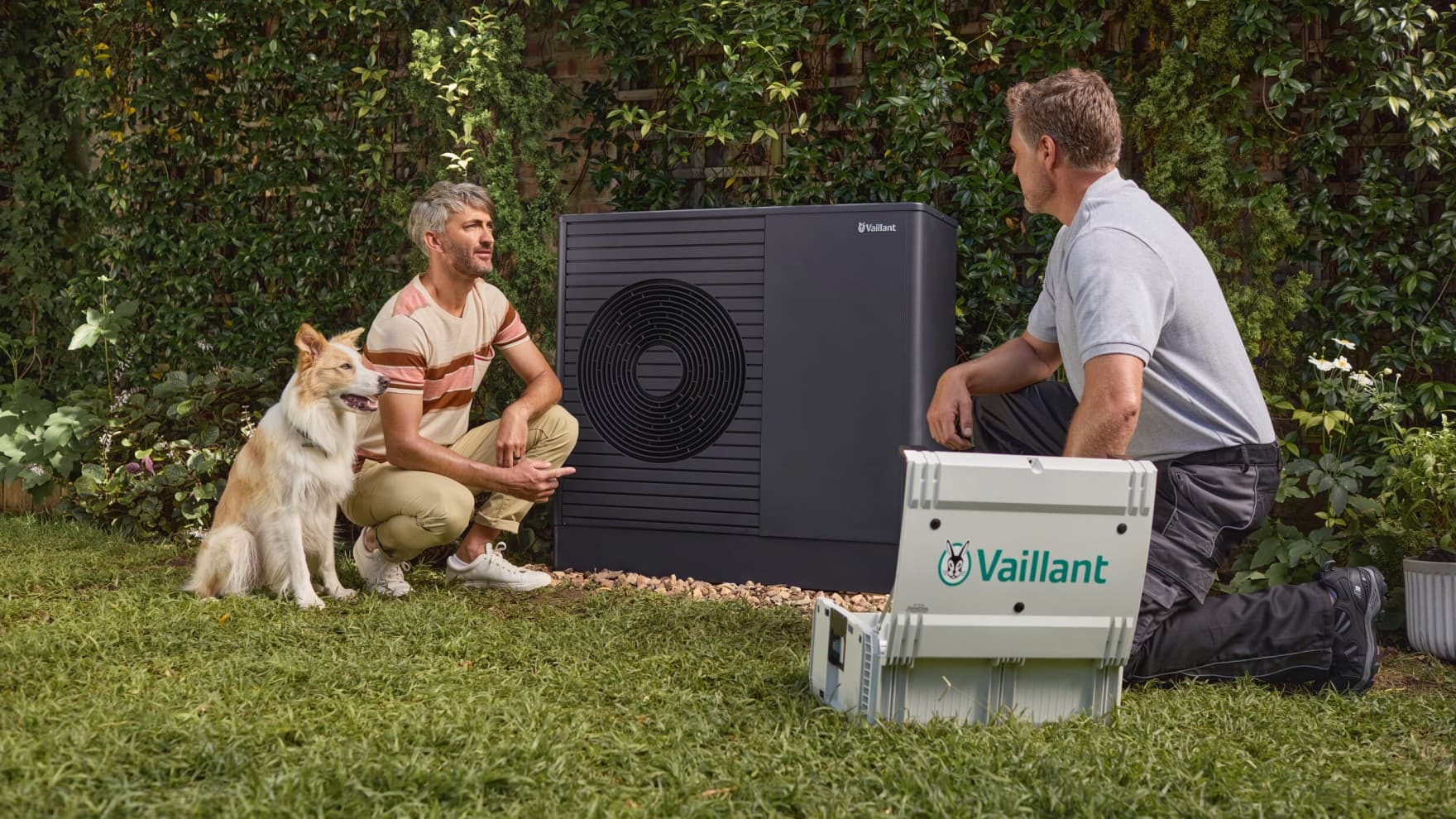 Eine Vaillant Wärmepumpe steht im Garten. Links daneben kniet der Eigentümer und sein Hund, rechts neben der Wärmepumpe der Heizungsfachmann.