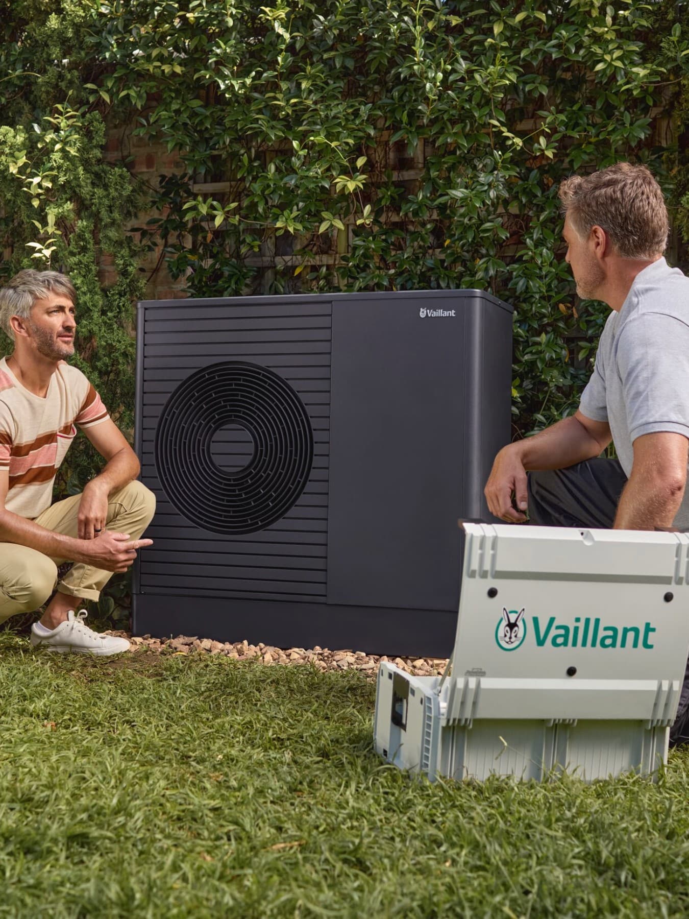 Eine Vaillant Wärmepumpe steht im Garten. Links daneben kniet der Eigentümer und sein Hund, rechts neben der Wärmepumpe der Heizungsfachmann.