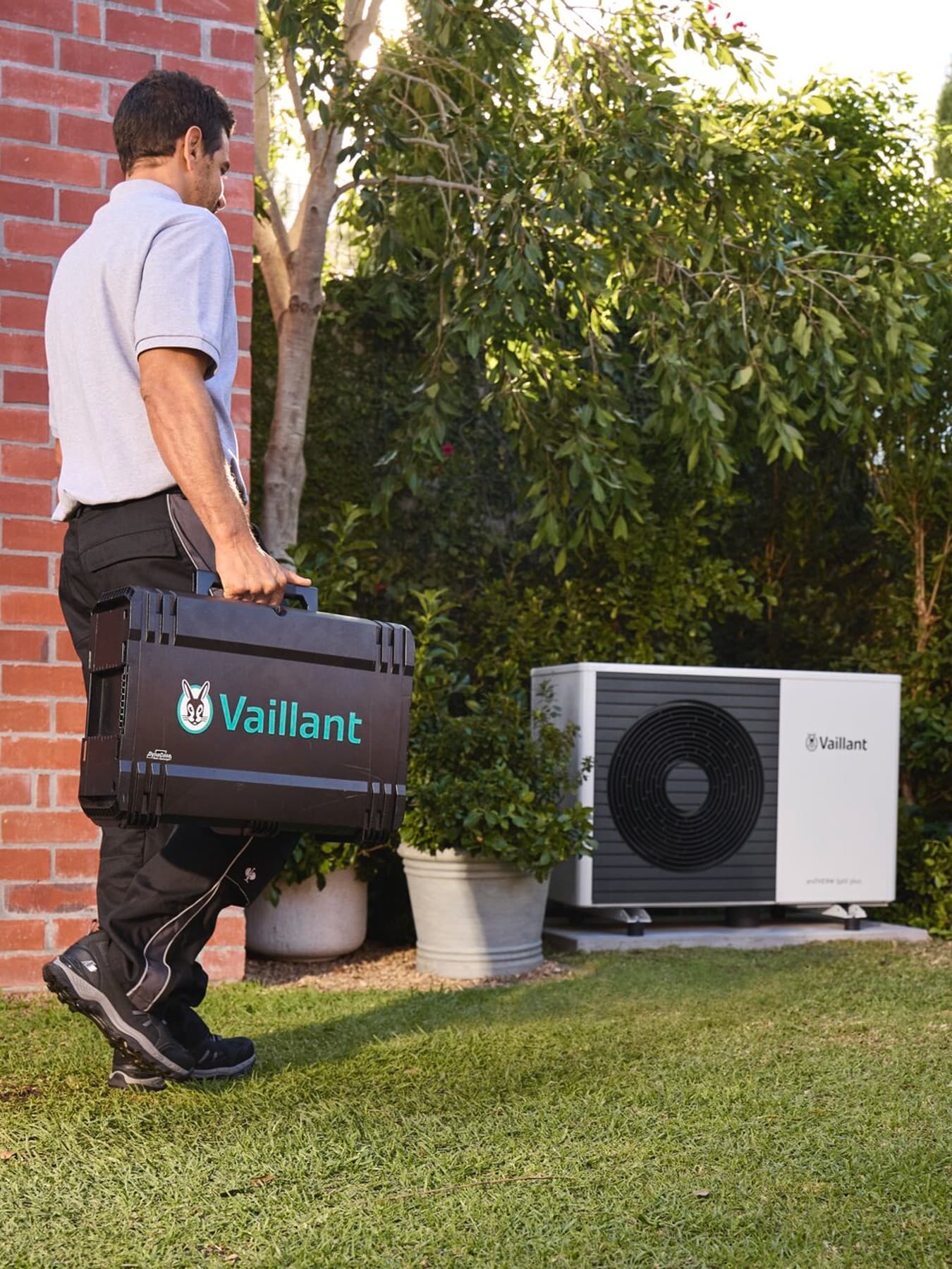 Techniker mit Vaillant-Werkzeugkoffer geht über eine Rasenfläche auf eine im Garten installierte Außeneinheit einer Wärmepumpe zu