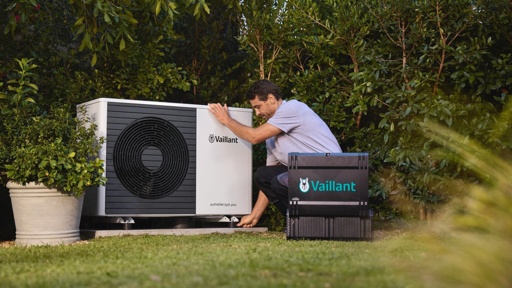 Ein Vaillant Techniker arbeitet an einer Vaillant Wärmepumpe, die in einem Garten vor einer Hecke steht; neben ihm steht ein Werkzeugkoffer.