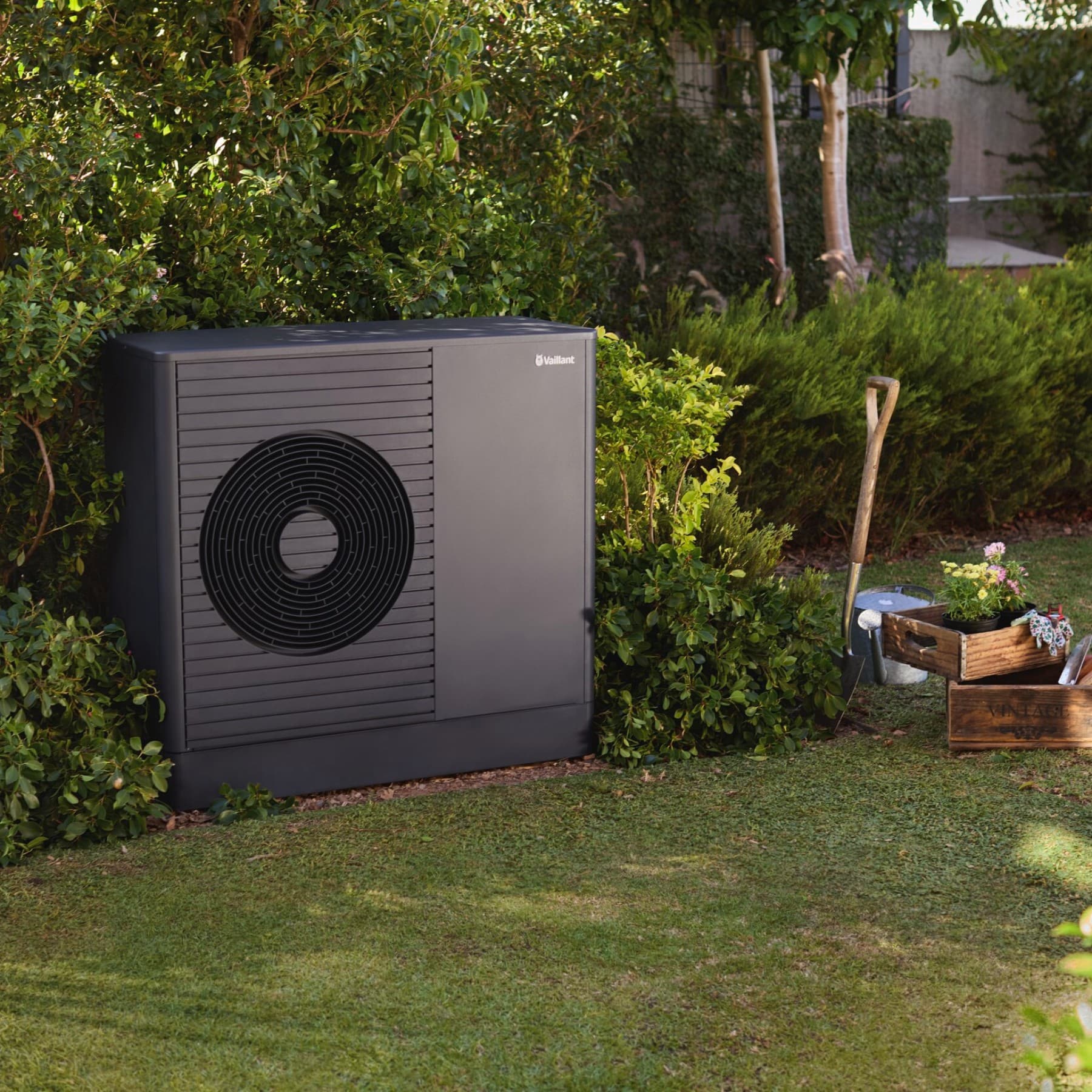 Vaillant Wärmepumpe im Garten, daneben ein Spaten und 2 Holkisten mit Blumen