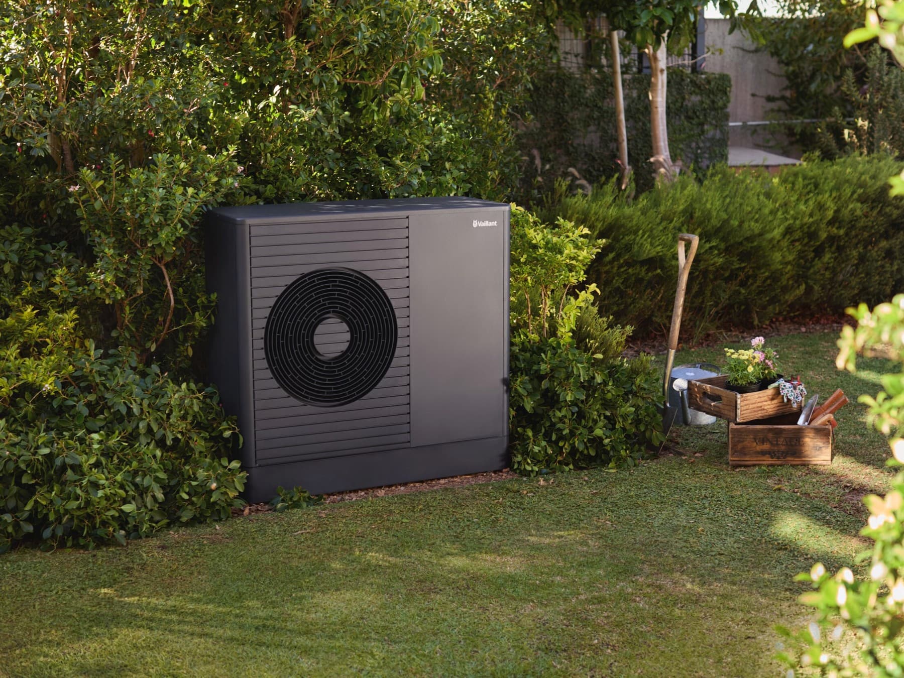 Vaillant Wärmepumpe im Garten, daneben ein Spaten und 2 Holkisten mit Blumen