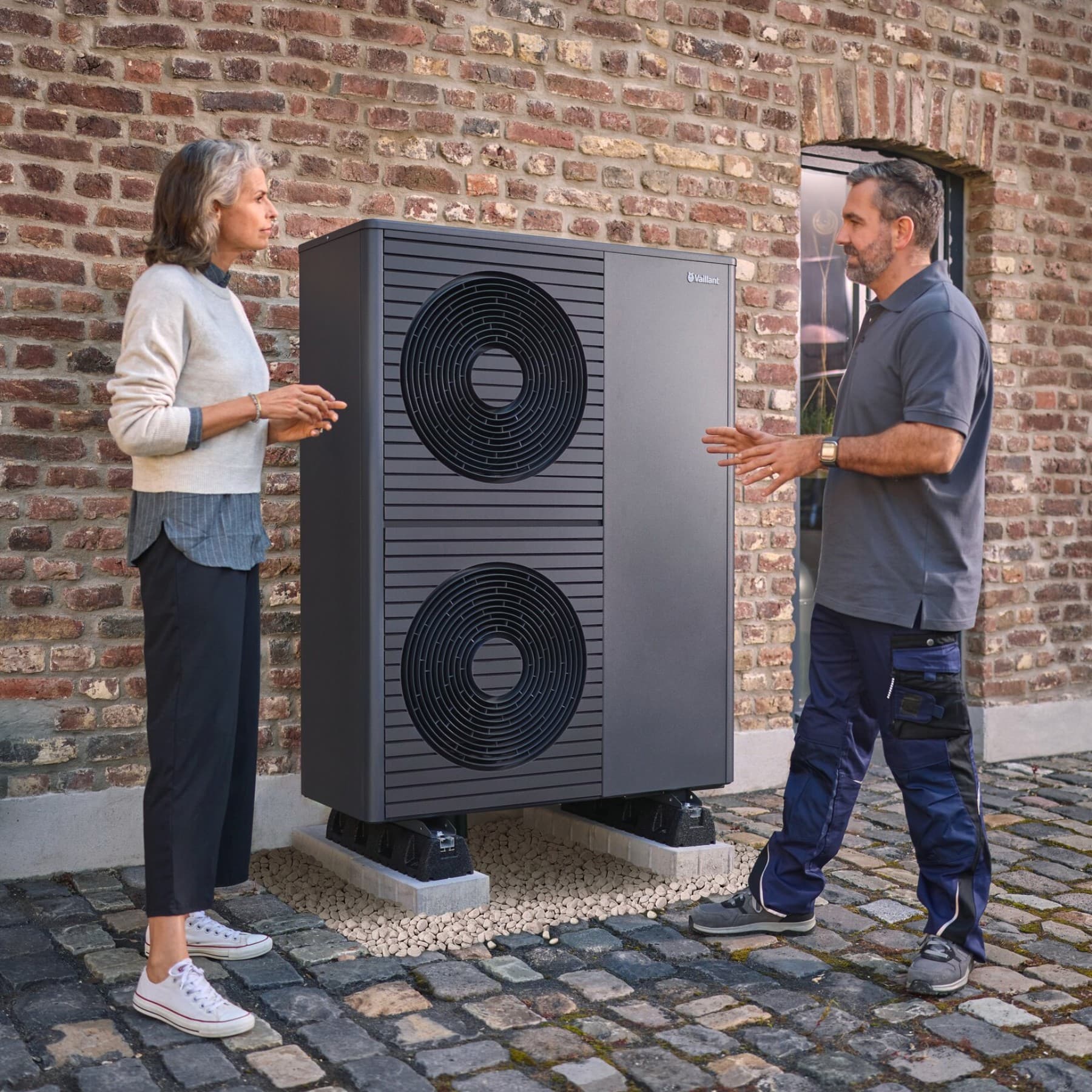Ein Vaillant Fachhandwerker steht links im Bild und erklärt einer Frau die Funktion einer Wärmepumpe. Zwischen ihnen steht vor einer Backstein-Hauswand eine Vaillant Wärmepumpe mit zwei Ventilatoren.