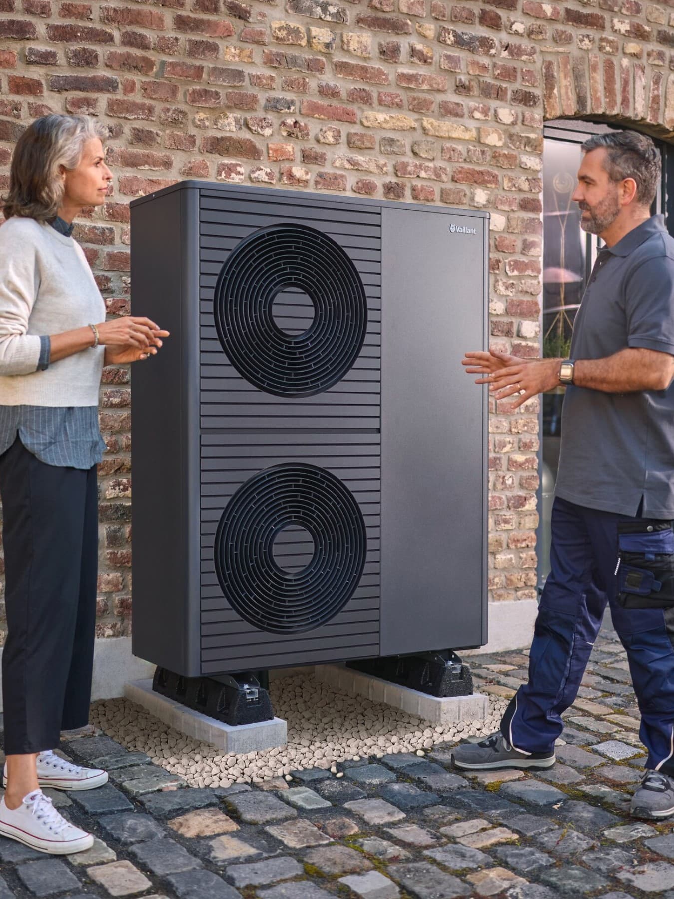 Fachhanwerker und Kundin stehen vor einer neu installierten aroTHERM plus Wärmepumpe vor einem Backsteinhaus