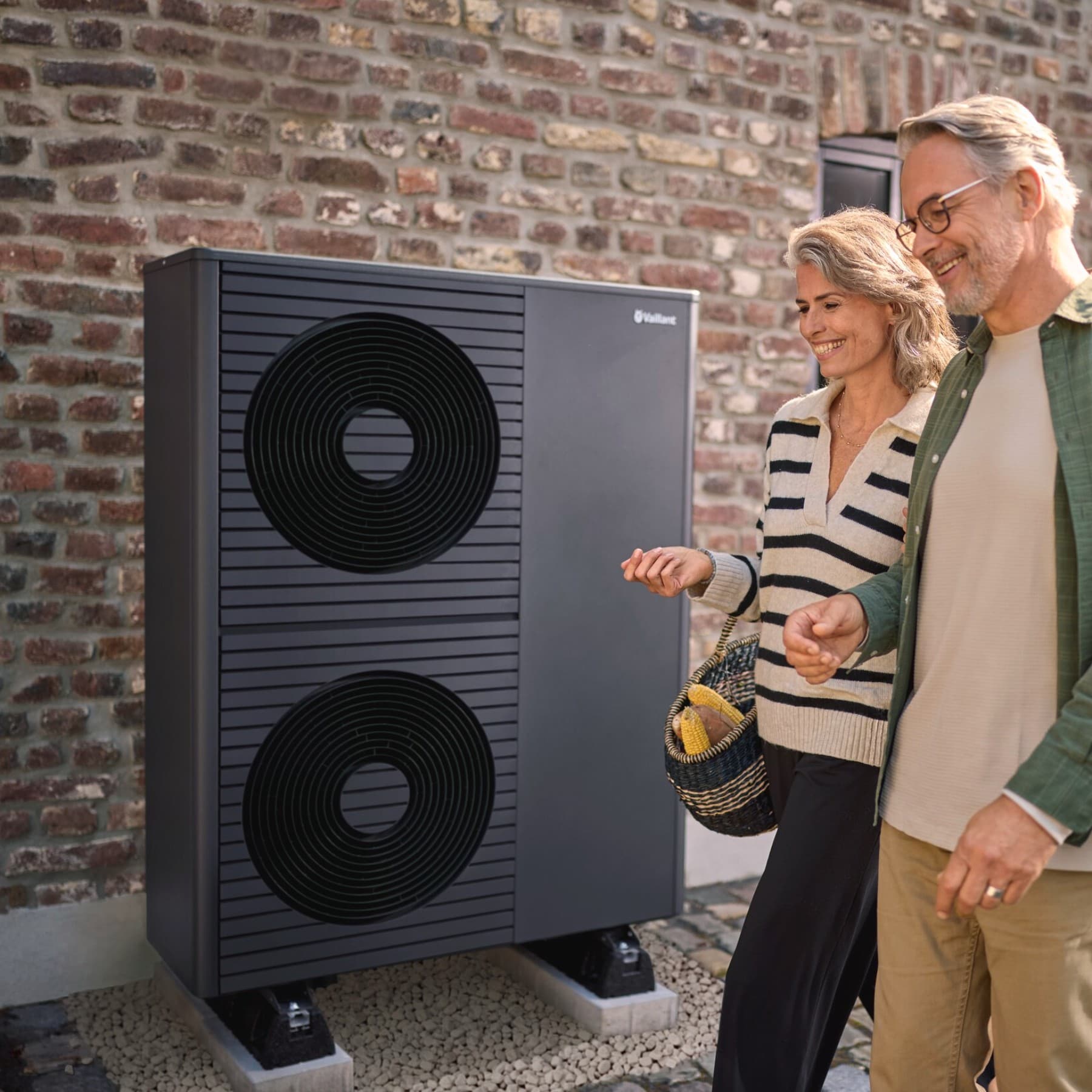 Ein Mann und eine Frau mit weiß-grauen Haaren gehen lächelnd an einem Backsteingebäude vorbei, vor dem eine Vaillant Wärmepumpe steht.