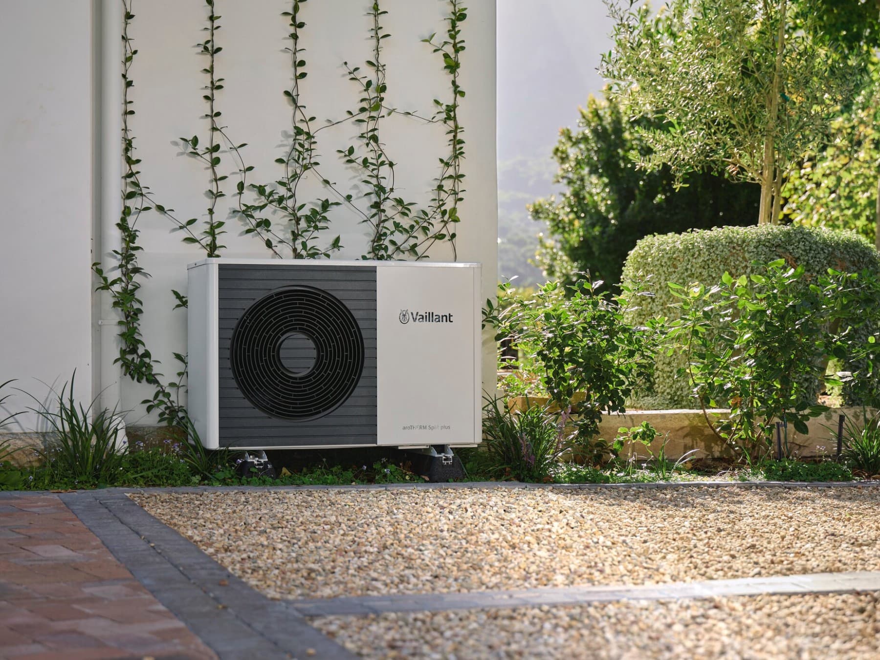 Eine Vaillant Wärmepumpe vor einer weißen Steinwand, umgeben von grünen Büschen und Pflanzen.