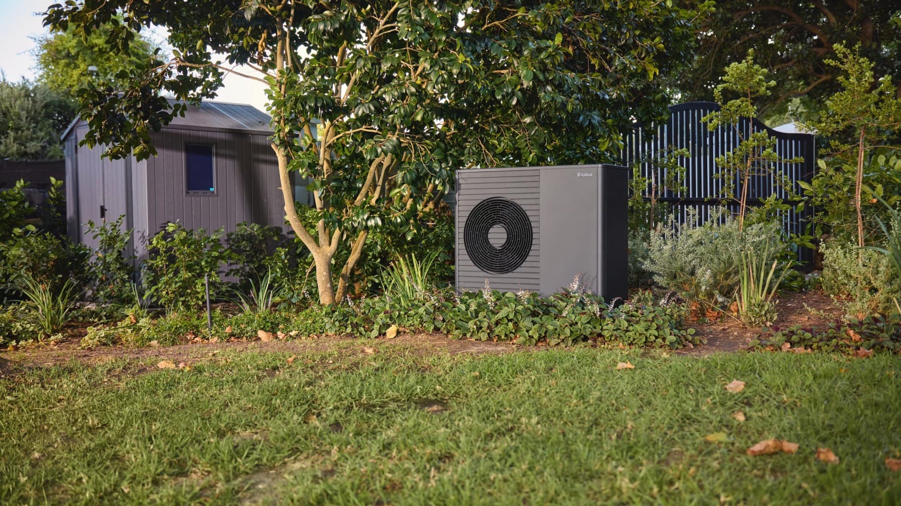 Eine Luft-Wasser-Wärmepumpe von Vaillant steht in einem Garten zwischen Büschen und Sträuchern, links daneben steht im Hintergrund ein Gartenhäuschen, hinter der Wärmepumpe ist ein Zaun zu sehen.