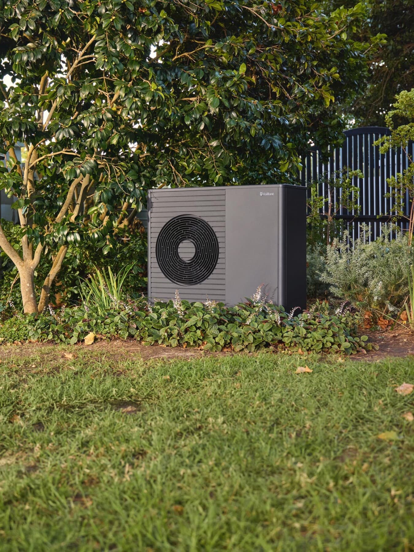Eine Luft-Wasser-Wärmepumpe von Vaillant steht in einem Garten zwischen Büschen und Sträuchern, links daneben steht im Hintergrund ein Gartenhäuschen, hinter der Wärmepumpe ist ein Zaun zu sehen.
