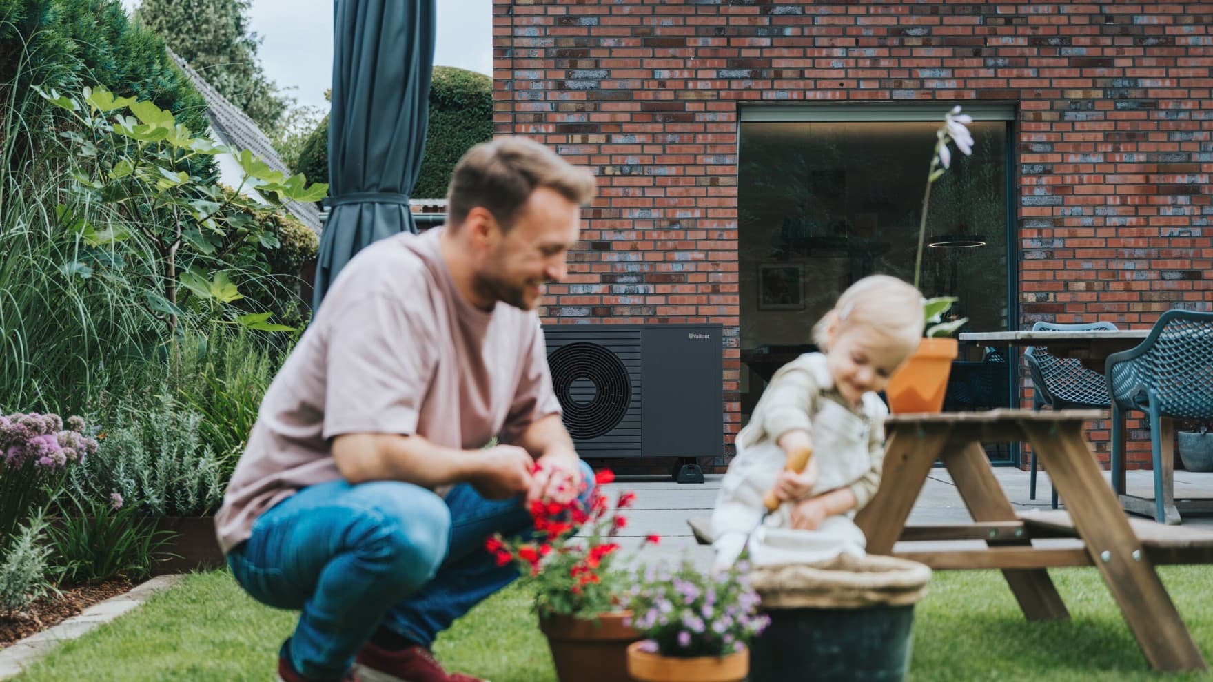 Vater und Tochter pflanzen Blumen in einen Blumentopf im Garten. Im Hintergrund sieht man ein Backsteinhaus. Auf der Terrasse steht eine Wärmepumpe aroTHERM plus.