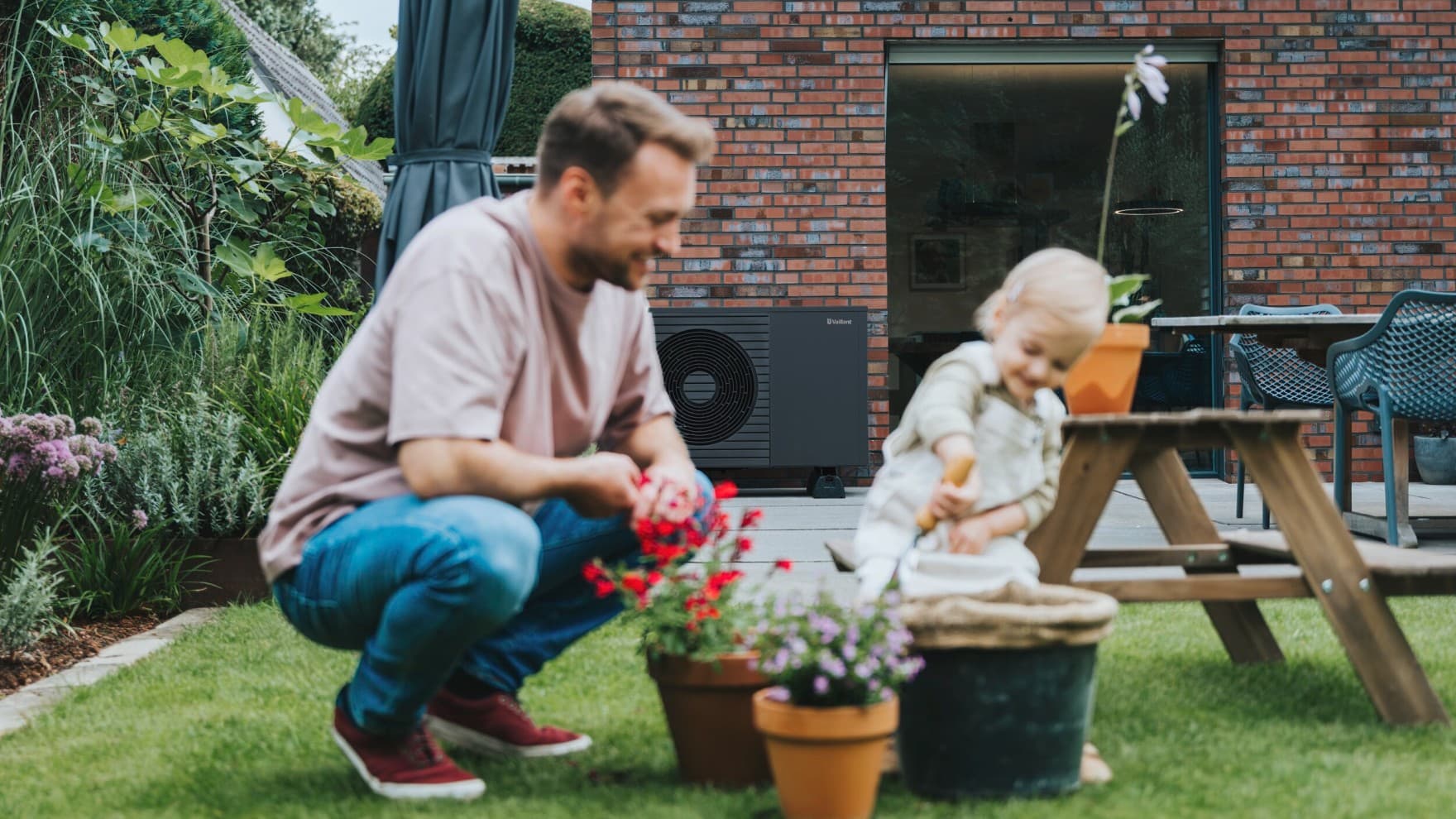 Kleines, lächelndes Mädchen sitzt auf einer aroTHERM plus Wärmepumpe im Garten mit Bäumen im Hintergrund