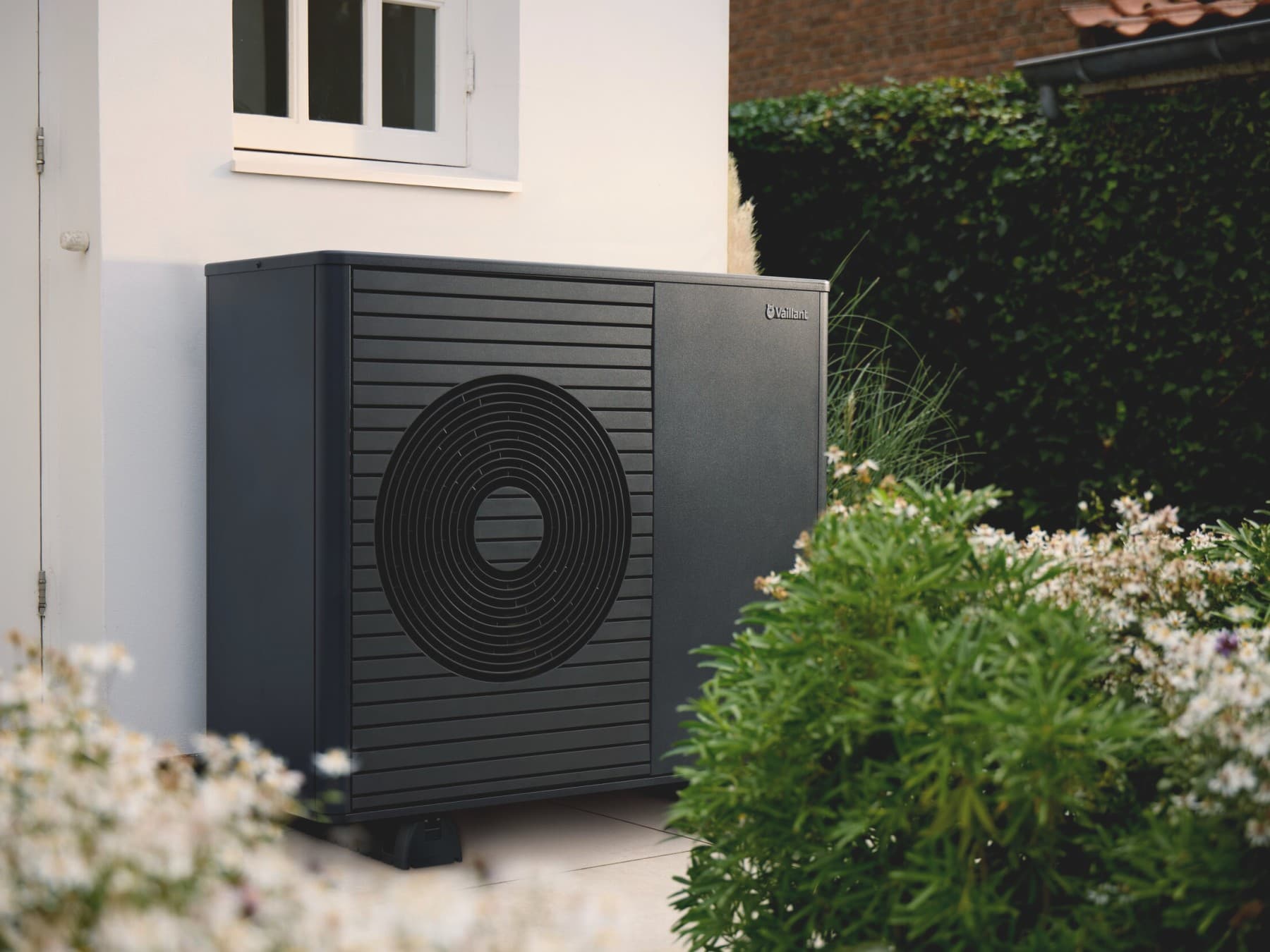 Die Vaillant Luft-Wasser-Wärmepumpe aroTHERM plus steht in einem Garten an einer weißen Hauswand zwischen Büschen und Beeten.