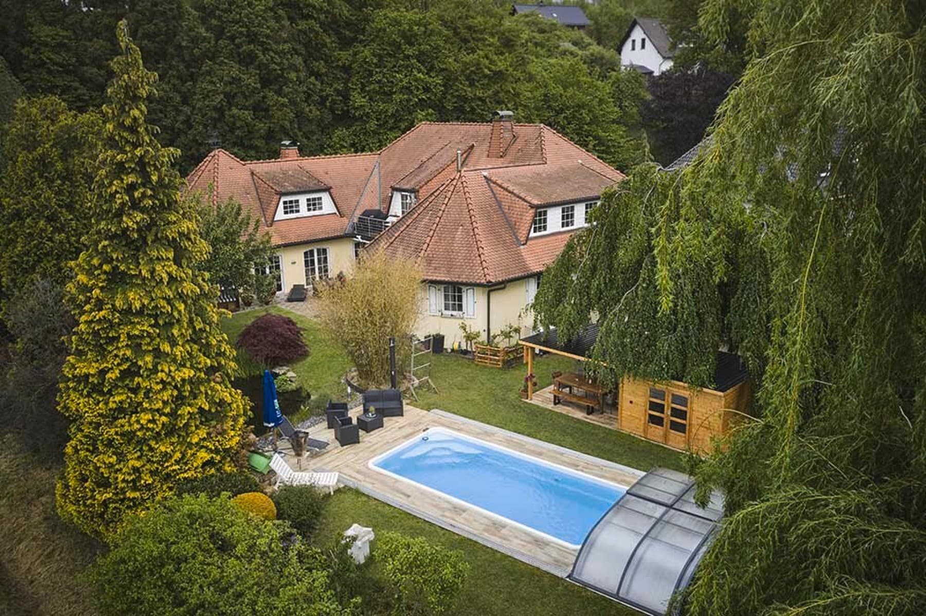 Das Haus in Porta Westfalica in Luftaufnahme. Zusehen ist ein Haus in U-Form, ein Garten mit Pool und ein großes Gartenhaus. Drumherum stehen viele Bäume.