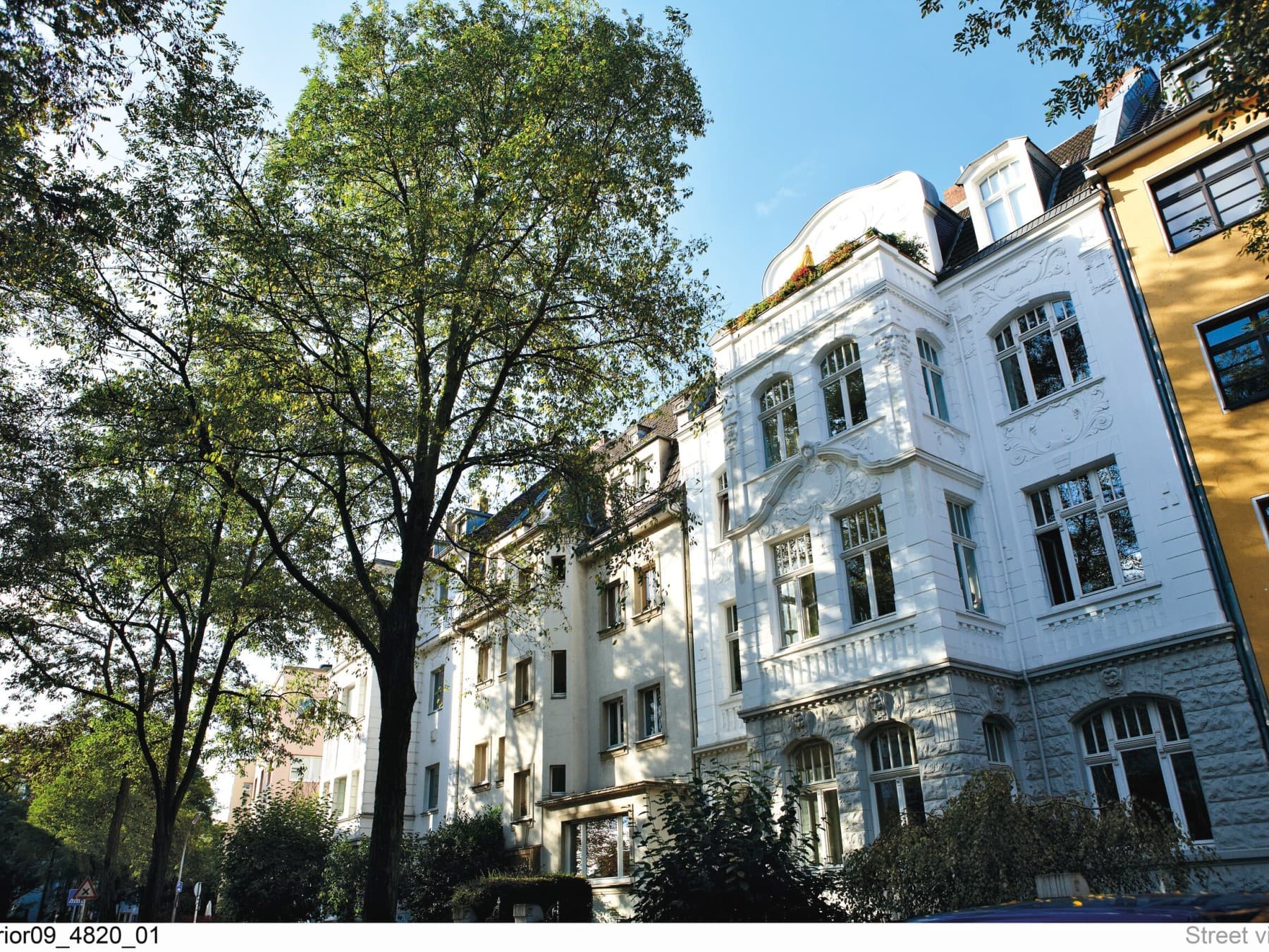 Straßenzug mit Mehrfamilienhäusern, Bäumen und blauem Sommerhimmel